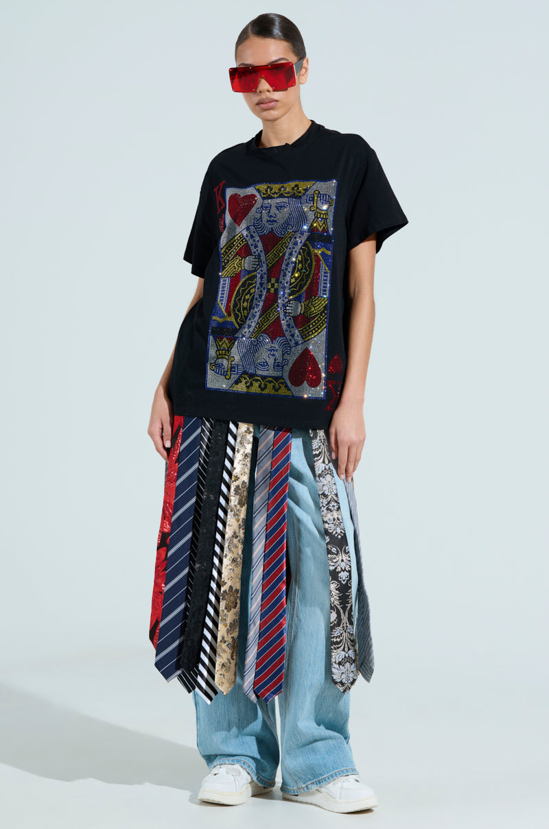 Wearing rhinestone graphic red sunglasses, a person matches their KING OF HEARTS OVERSIZED TEE with loose blue jeans and an eclectic necktie skirt. They pose confidently against a plain light blue backdrop.