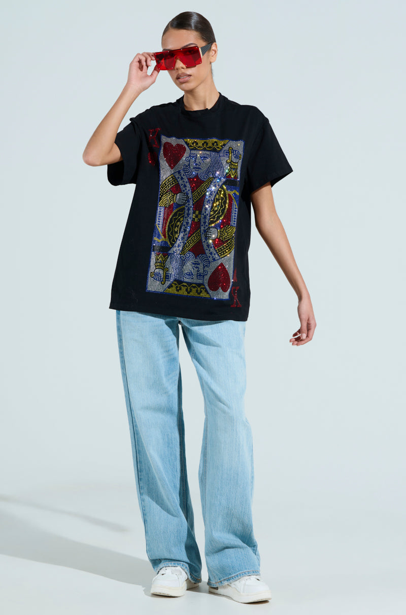 Wearing red sunglasses, a KING OF HEARTS OVERSIZED TEE, light blue wide-leg jeans, and white sneakers against a plain background, they hold the sunglasses with one hand, showcasing their effortlessly cool style.