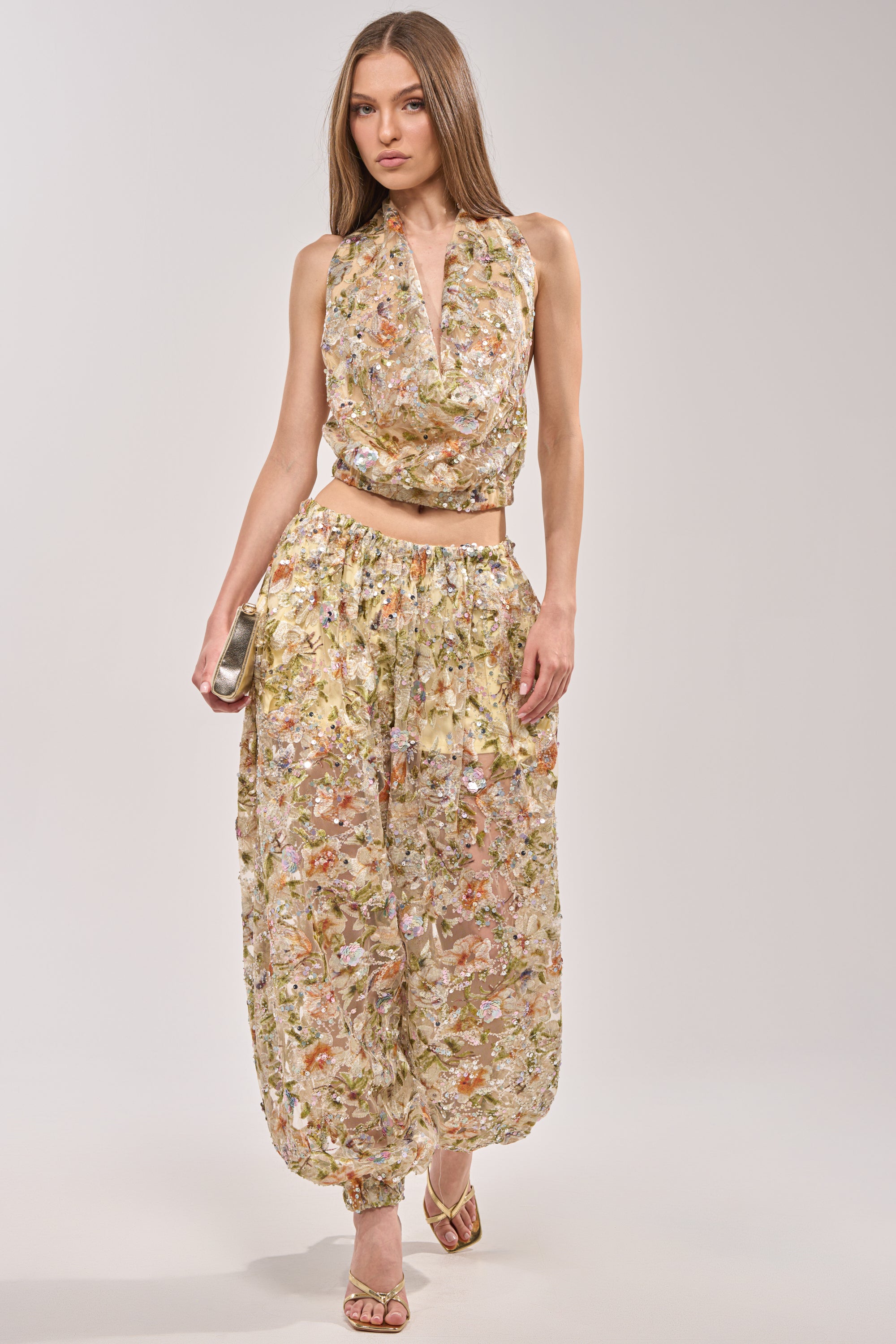 A woman stands against a plain light background wearing the LETS TALK ABOUT IT COWL NECK top with matching sheer, flowy pants, heeled sandals, and holding a small clutch. 9