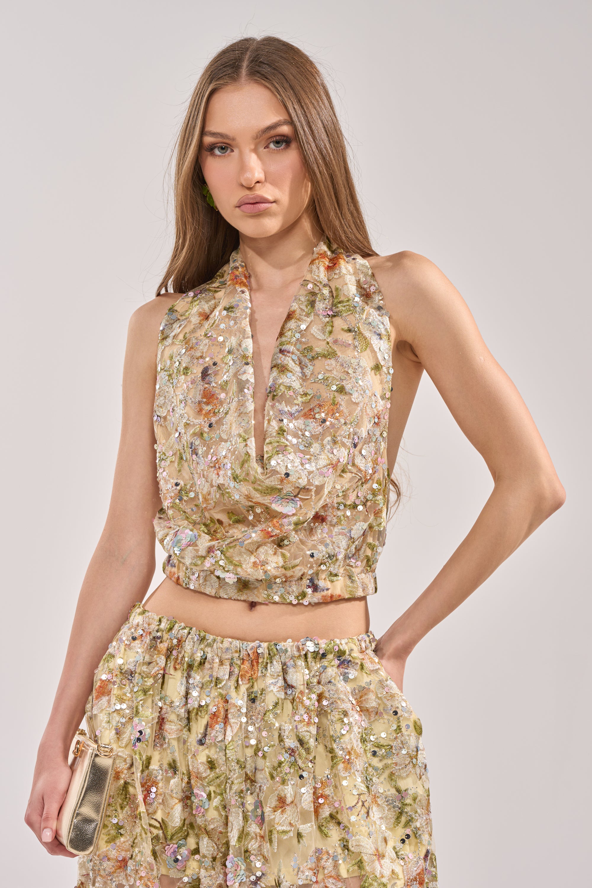 A woman with long brown hair wears the LETS TALK ABOUT IT COWL NECK floral embroidered mesh crop top and matching high-waisted skirt, holding a small metallic clutch against a light gray background. 3
