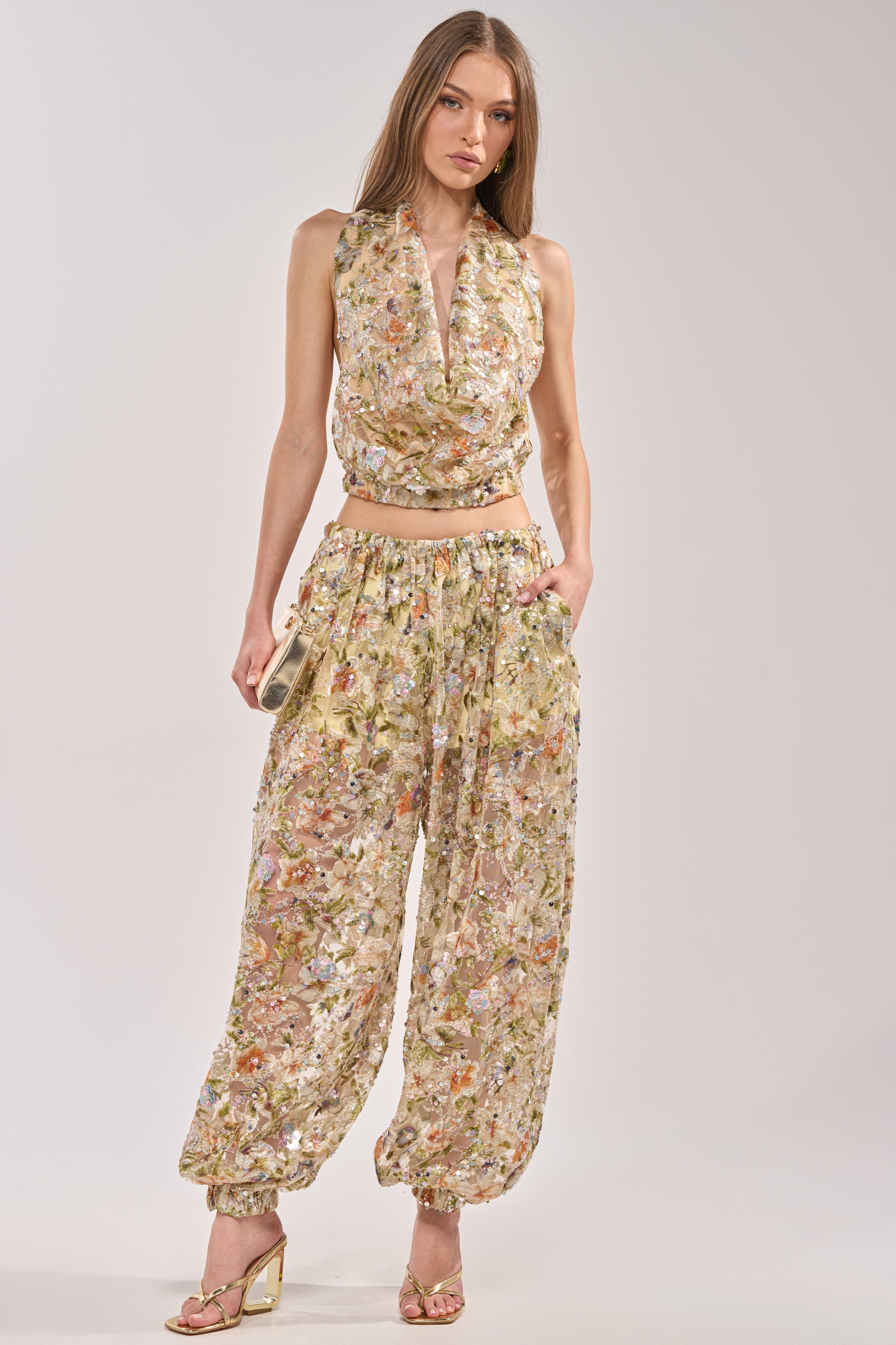 A woman poses with one hand in her pocket against a plain background, wearing the LETS TALK ABOUT IT COWL NECK sleeveless floral top, matching high-waisted pants, and gold high-heeled sandals. 6