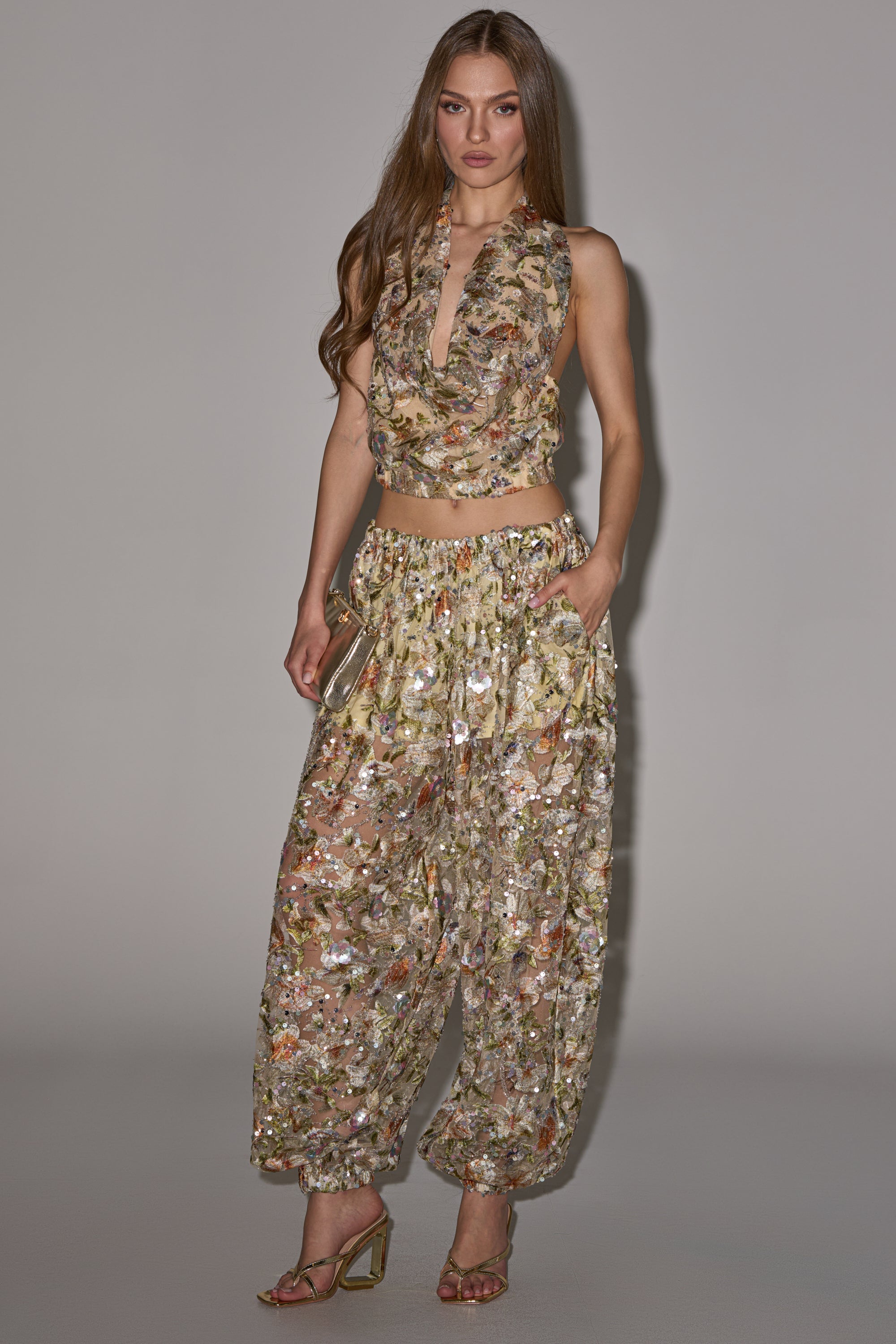 A woman poses against a plain background wearing the LETS TALK ABOUT IT COWL NECK top with wide-leg pants. She has long hair, carries a small clutch, and finishes her look with strappy heels. 7