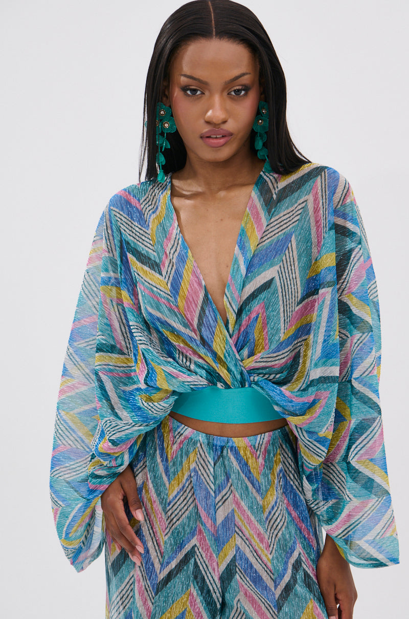 A woman with long straight hair wears large green earrings and the GRISELDA PRINTED CROP LONG SLEEVE TOP, which features a deep V-neck, batwing sleeves, turquoise waistband, and a colorful geometric chevron print in blue, yellow, pink, and green.
