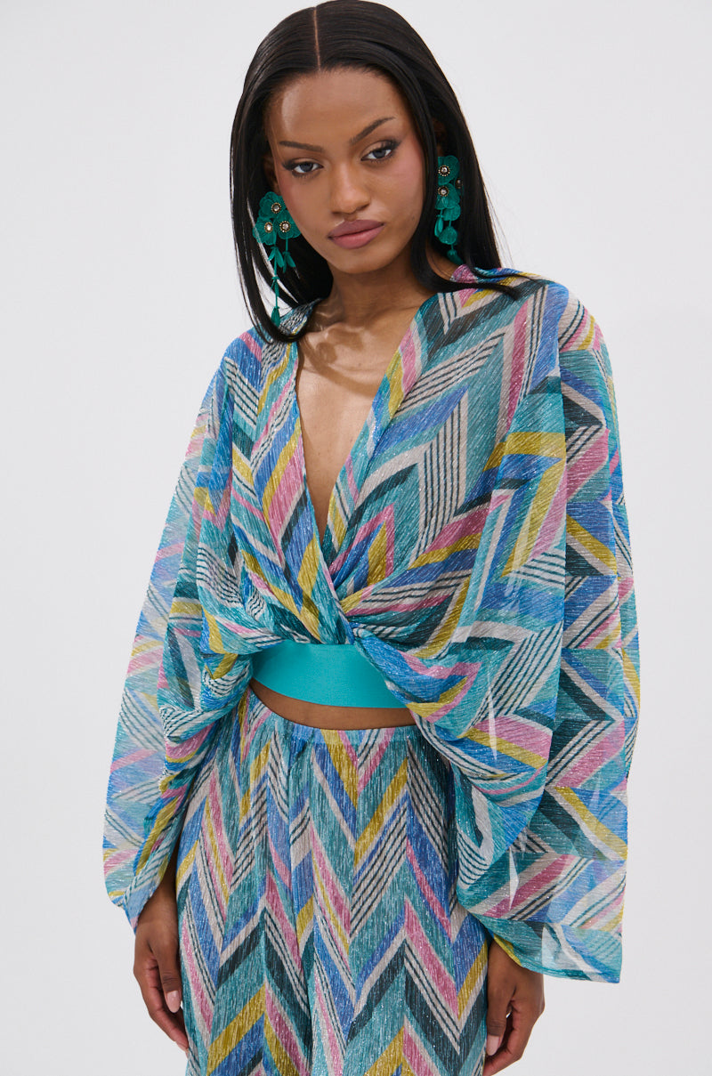 A woman models the GRISELDA PRINTED CROP LONG SLEEVE TOP with a geometric multicolor pattern, batwing sleeves, and teal waistband. She wears bold green earrings and has long straight hair against a white background.