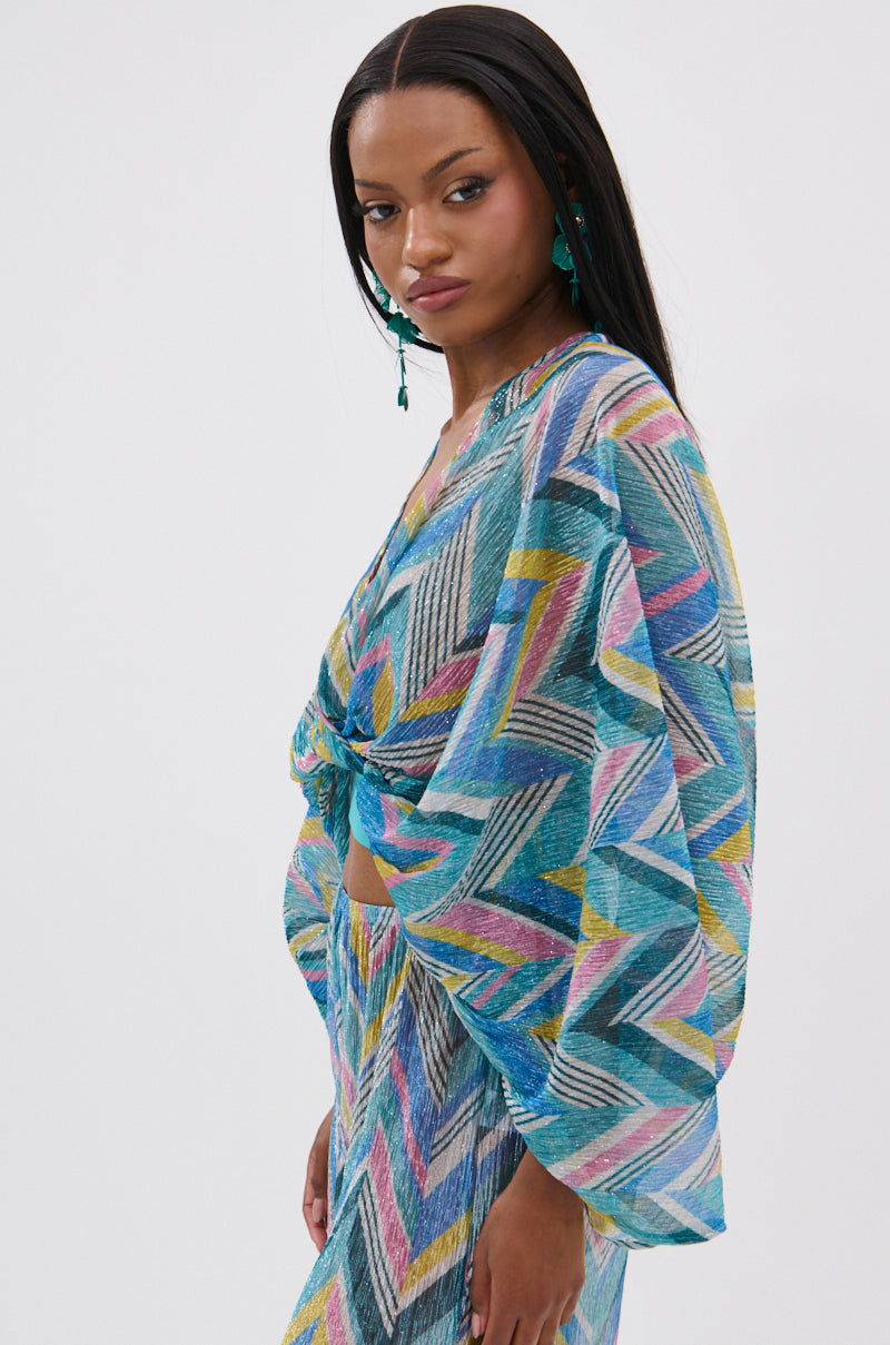 A woman with long straight hair wears the GRISELDA PRINTED CROP LONG SLEEVE TOP in a colorful geometric print with batwing sleeves and a front knot, standing in profile against a white background and accessorized with turquoise earrings.