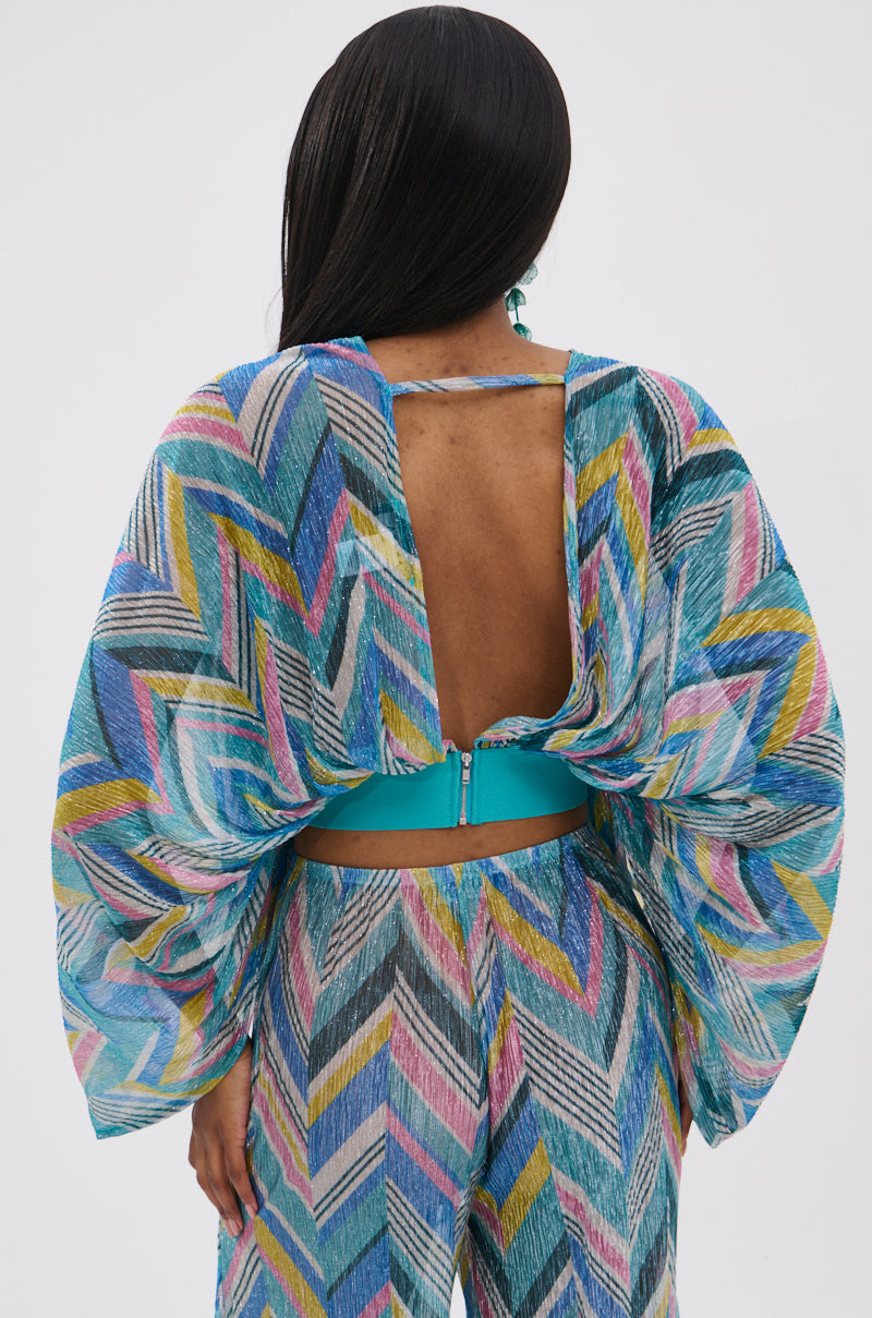 A person with long dark hair is shown from the back, wearing the GRISELDA PRINTED CROP LONG SLEEVE TOP with a geometric multicolor print, voluminous batwing sleeves, and an open back secured by a blue belt and zipper against a plain light background.