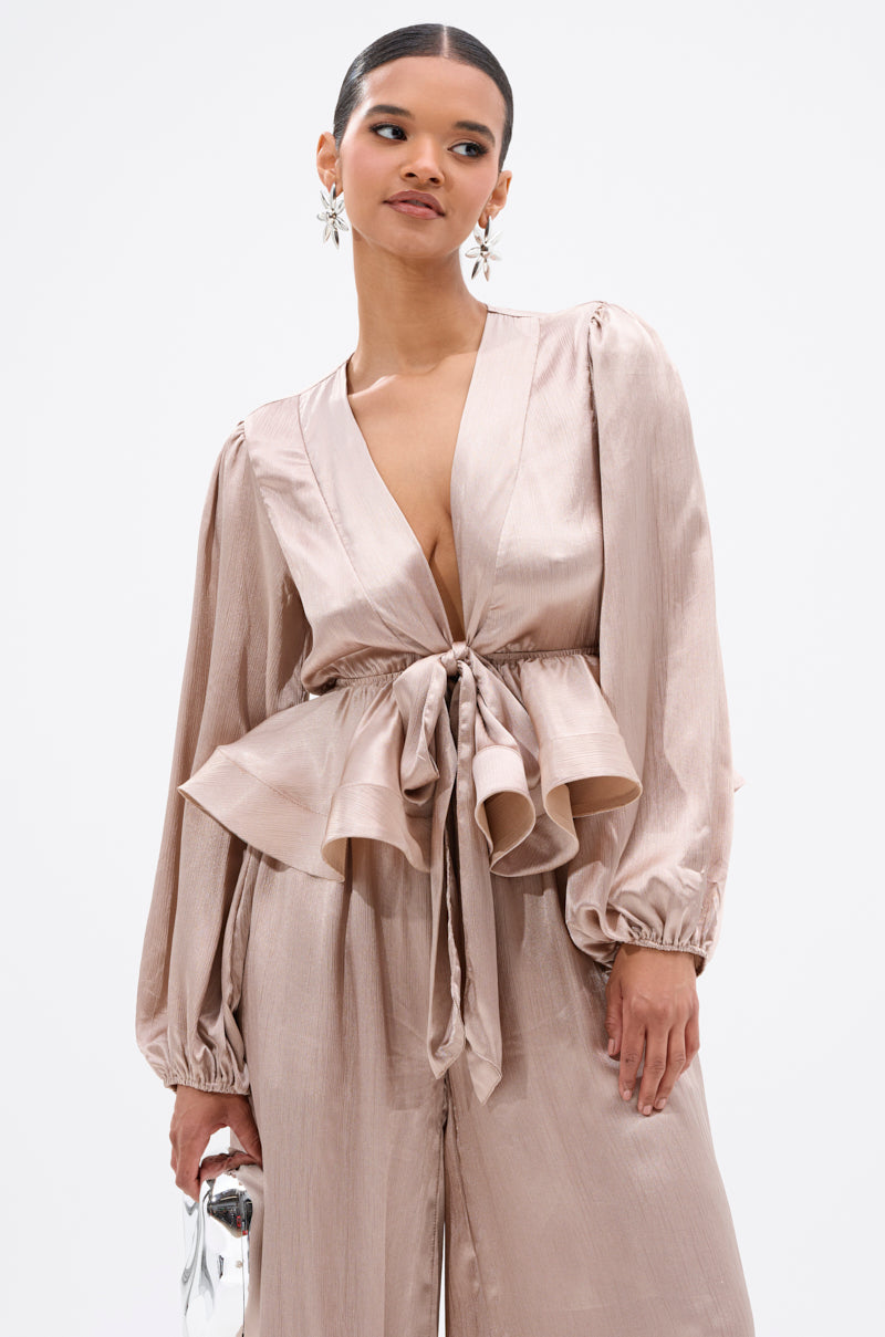 A woman in the ELEGANT PEPLUM TIE FRONT BLOUSE—shiny light taupe with a deep V-neck, long puffed sleeves, and peplum waist—stands against a white background holding a small silver clutch. 0