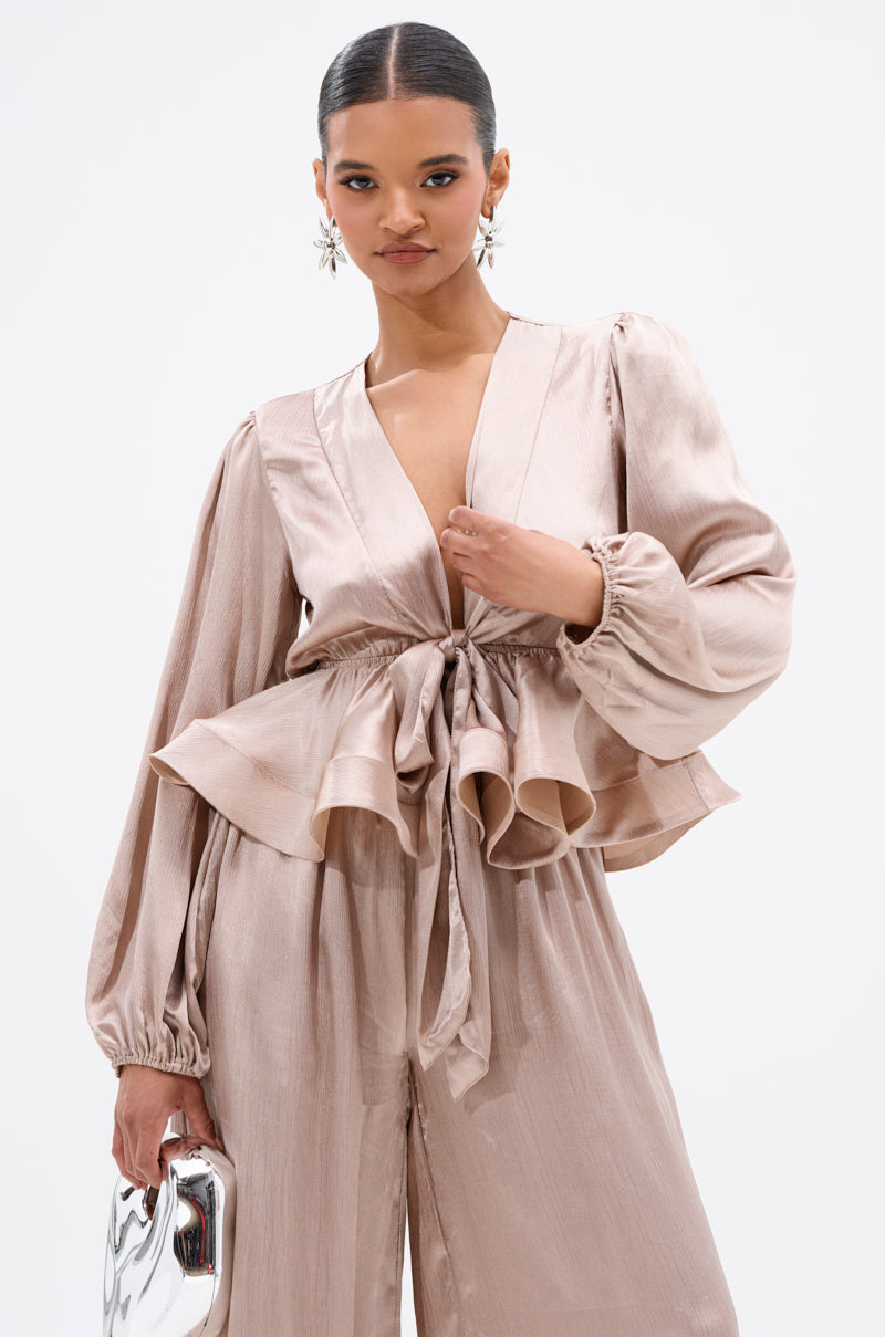 A woman with slicked-back hair wears the ELEGANT PEPLUM TIE FRONT BLOUSE in light beige, featuring a deep V-neck, long puffed sleeves, and a tied waist, paired with matching wide-leg pants. She carries a silver clutch and wears statement earrings. 1