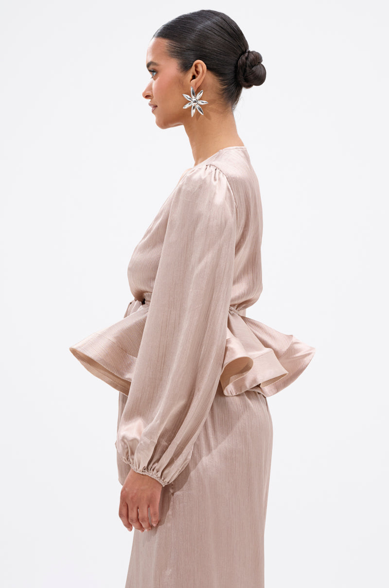 A woman in profile wears the ELEGANT PEPLUM TIE FRONT BLOUSE in pale pink with long sleeves and a matching skirt. Her hair is styled in a sleek bun, complemented by large star-shaped earrings against a plain white background. 2