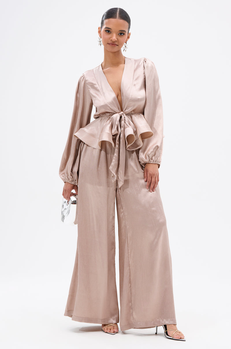 A woman wearing the ELEGANT PEPLUM TIE FRONT BLOUSE in blush with a wide sash belt and matching wide-leg pants, holding a small silver bag, stands against a plain white background. 4
