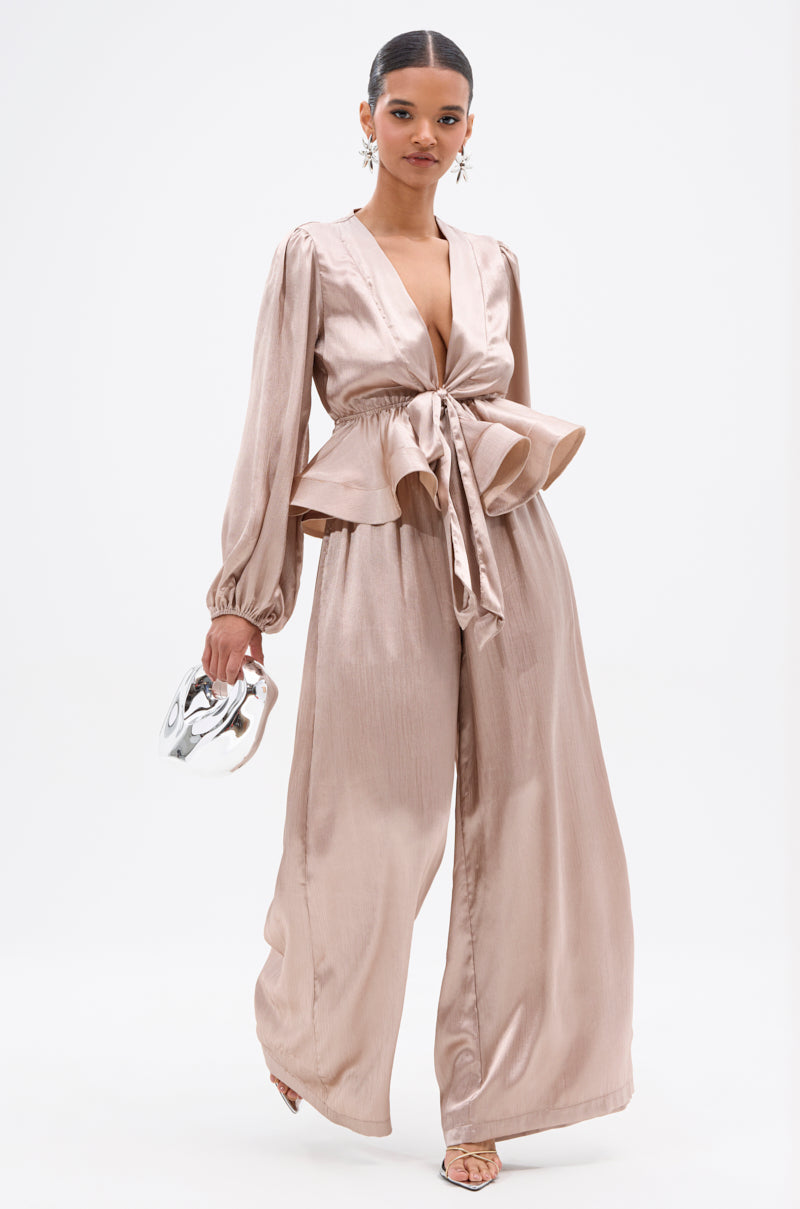 A woman stands confidently wearing the ELEGANT PEPLUM TIE FRONT BLOUSE in a shiny champagne hue, paired with wide-leg pants, statement earrings, sandals, and a metallic purse against a plain white background. 5