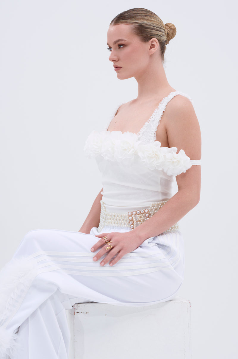 A woman with blonde hair in a sleek bun wears the AURA FLORAL TANK TOP, featuring floral appliques, as she sits sideways on a white block against a plain light background.