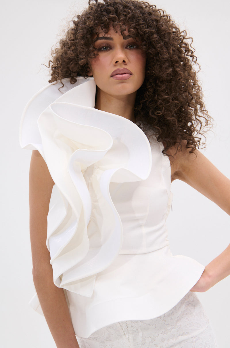 A woman with curly hair models the JULIETTE EXAGERATD RUFFLE BLOUSE, a chic white sleeveless top with dramatic ruffles on one shoulder, posing confidently against a simple light background.