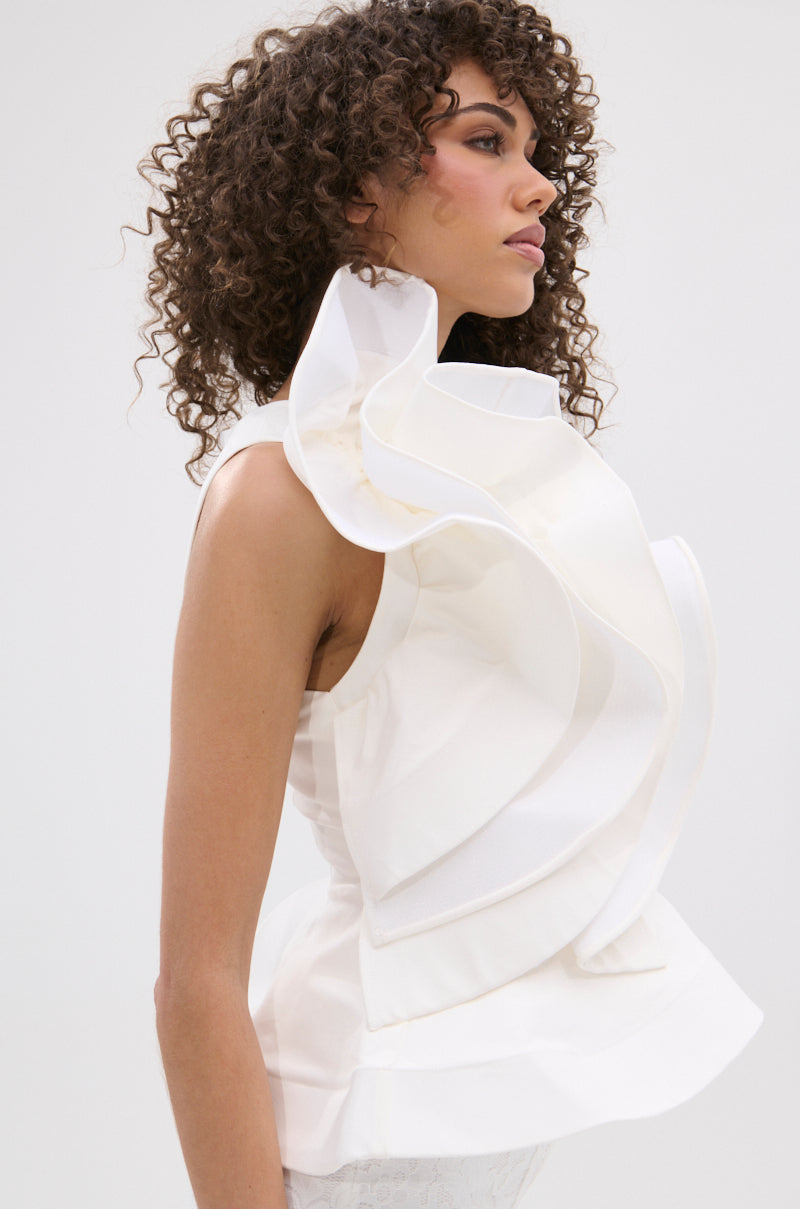 A woman with curly hair models the JULIETTE EXAGERATD RUFFLE BLOUSE, a sleeveless white top featuring dramatic ruffles at the neckline and shoulders, while posing in profile against a plain light background.