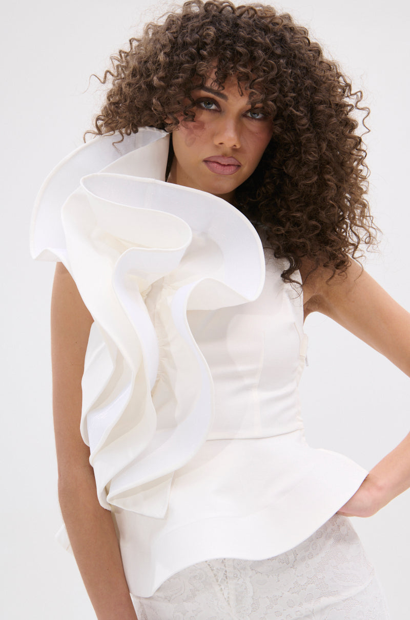 A curly-haired woman wears the JULIETTE EXAGERATD RUFFLE BLOUSE, showcasing dramatic ruffles on one shoulder and a peplum silhouette, paired with white lace bottoms against a simple light background.