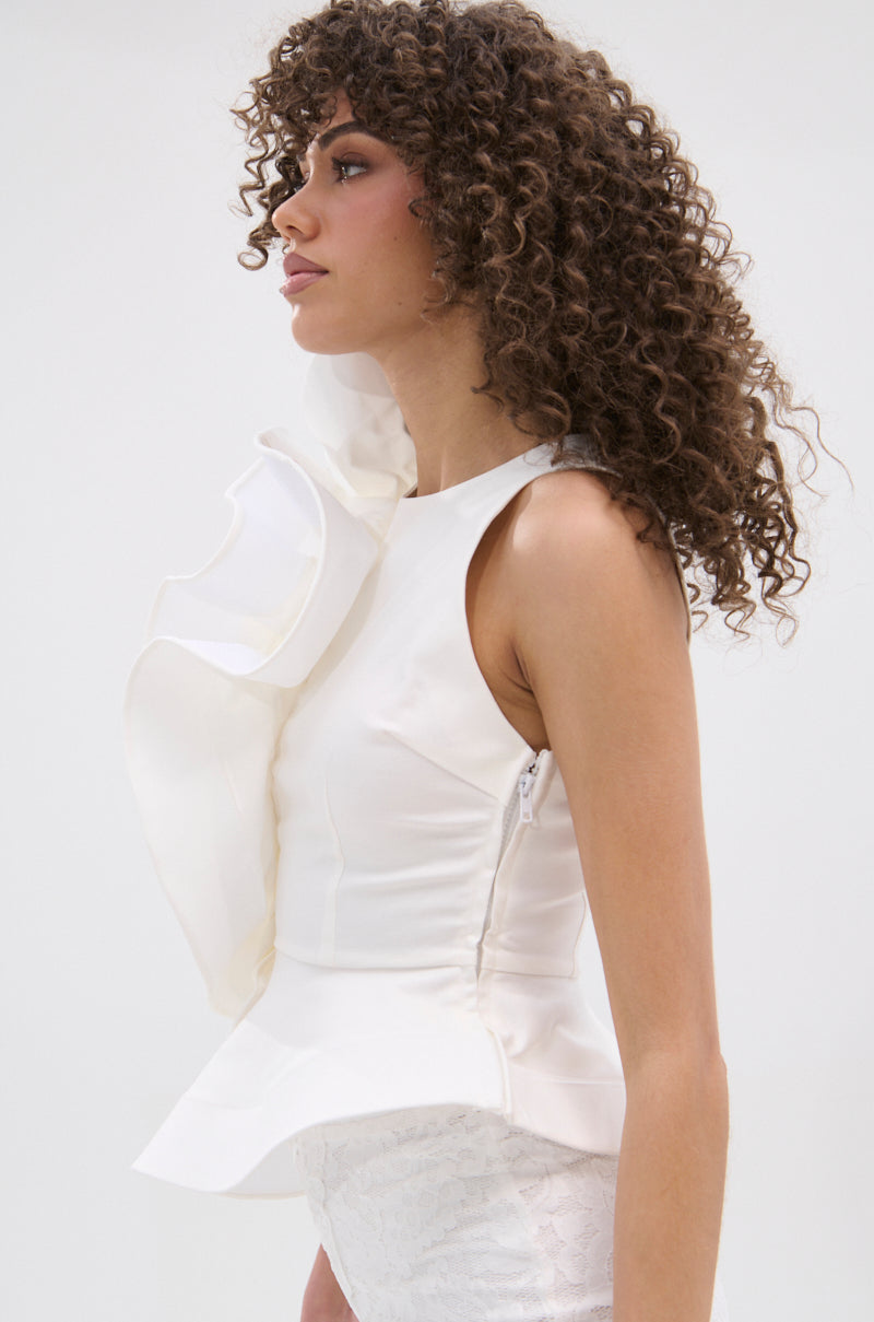 A woman with curly hair wears the JULIETTE EXAGERATD RUFFLE BLOUSE—a sleeveless, fitted white top with dramatic front ruffles—paired with white textured bottoms, standing against a plain light background.