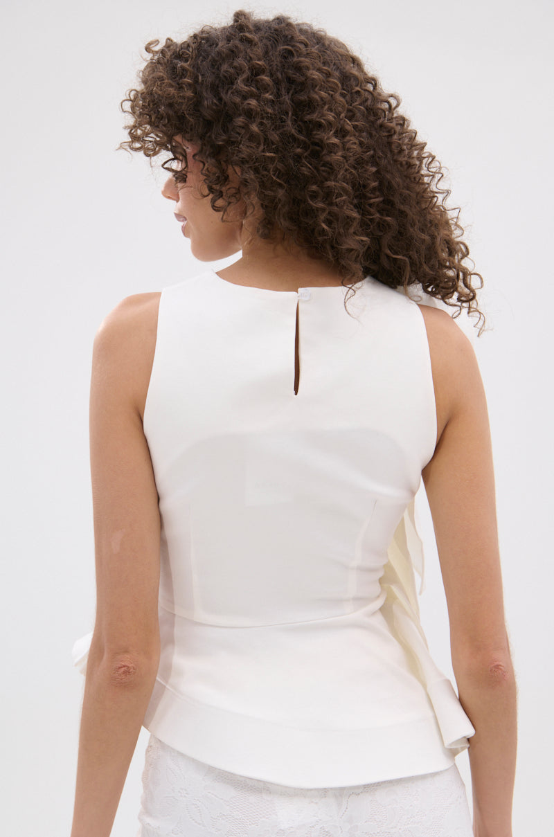 A woman with curly hair is seen from the back wearing the sleeveless Juliette Exaggerated Ruffle Blouse in white, featuring a fitted waist, subtle peplum silhouette, and a small button closure at the neck against a plain light background.