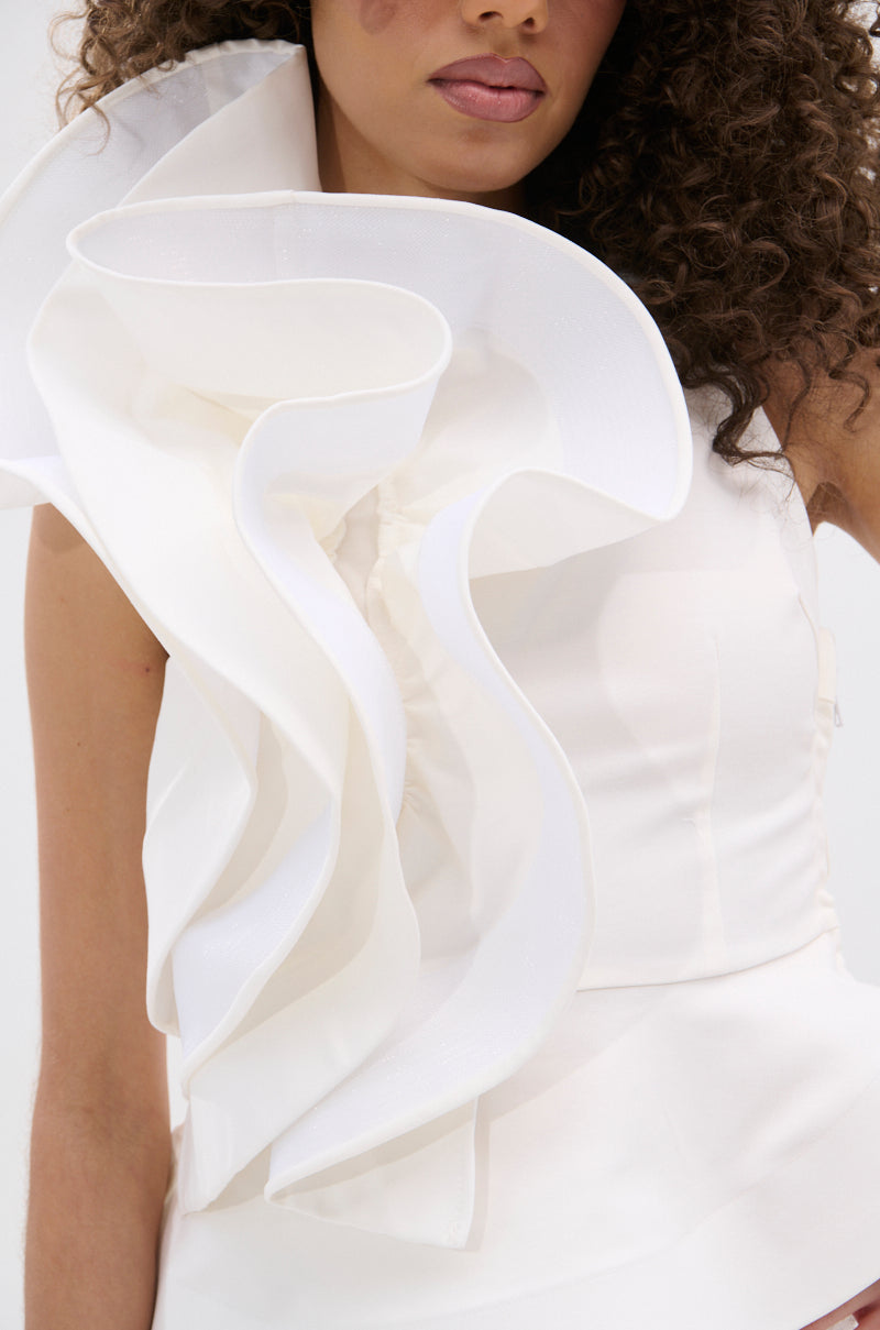 A woman with curly hair wears the JULIETTE EXAGERATD RUFFLE BLOUSE in white, featuring dramatic ruffles from her shoulder to chest. Only the upper part of her body is shown.