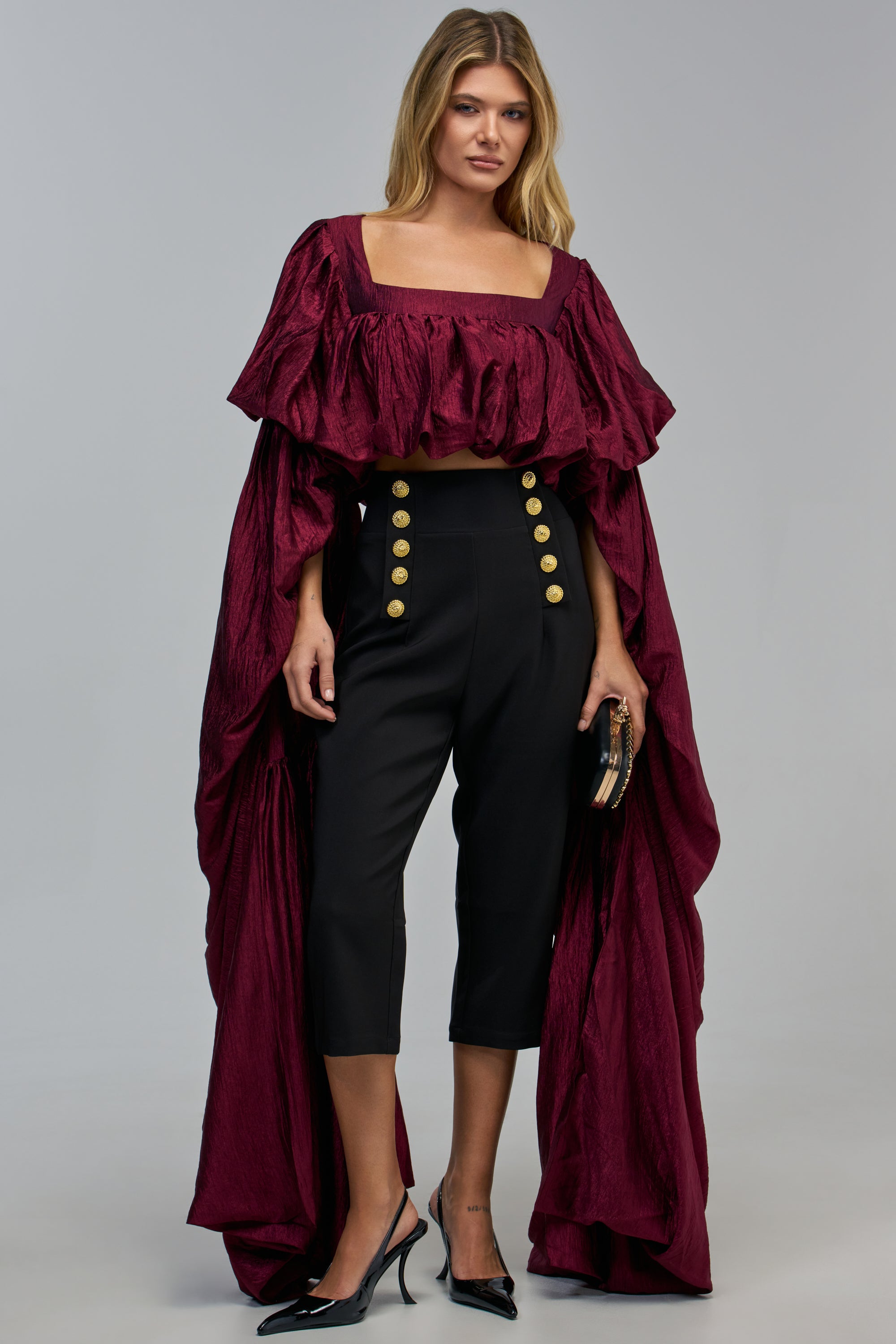 A model wears the GILDED EXCELLENCE TOP, a cropped, deep red burgundy satin blouse with ruffled off-the-shoulder draped sleeves and long back panels, paired with high-waisted black trousers featuring gold buttons.