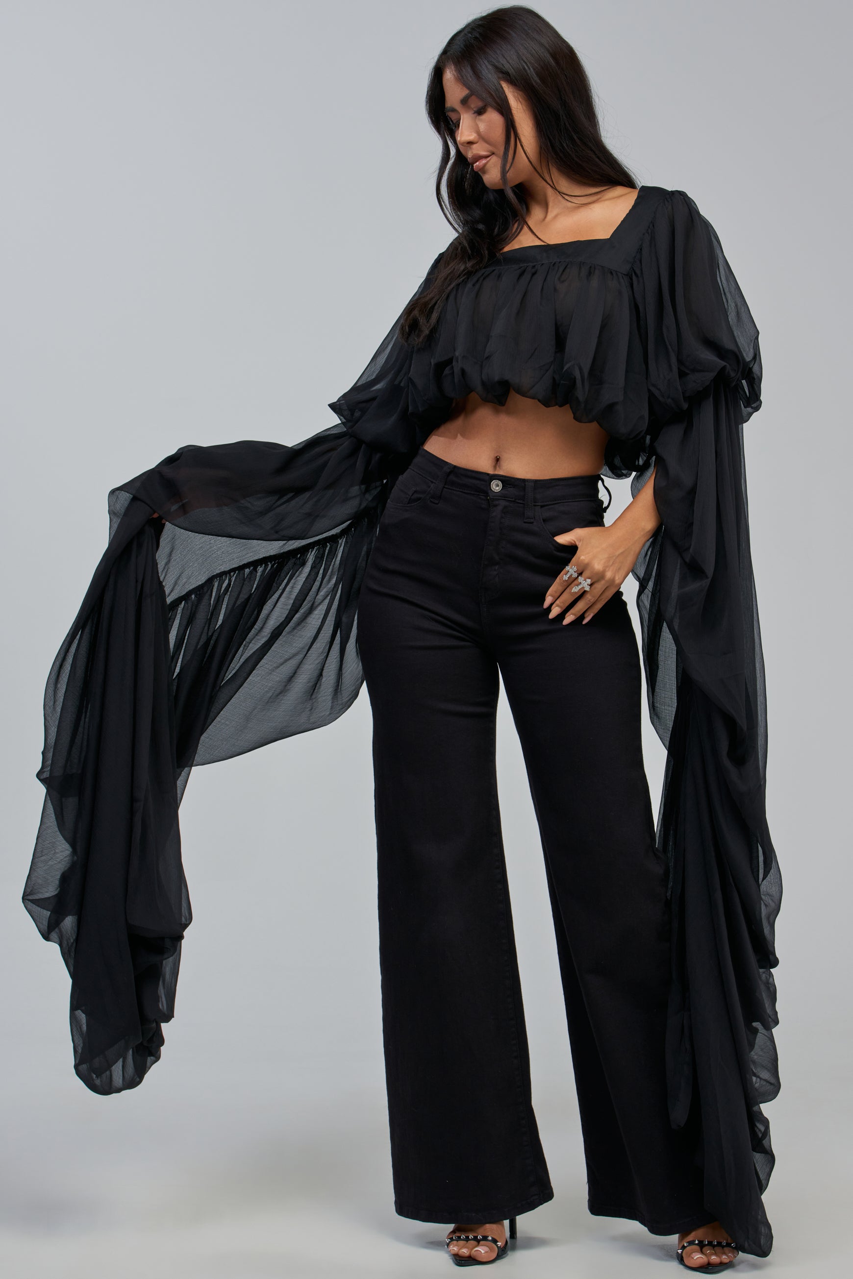 A woman stands smiling with her hand on her hip, wearing the SLOUCHY SQUARE NECK LONG SLEEVE BLOUSE IN BLACK styled as a crop top. The dramatic blouse features long, flowing sheer sleeves that drape to the floor. She pairs it with wide-leg black pants, and her long dark hair cascades over her shoulders against a plain background.