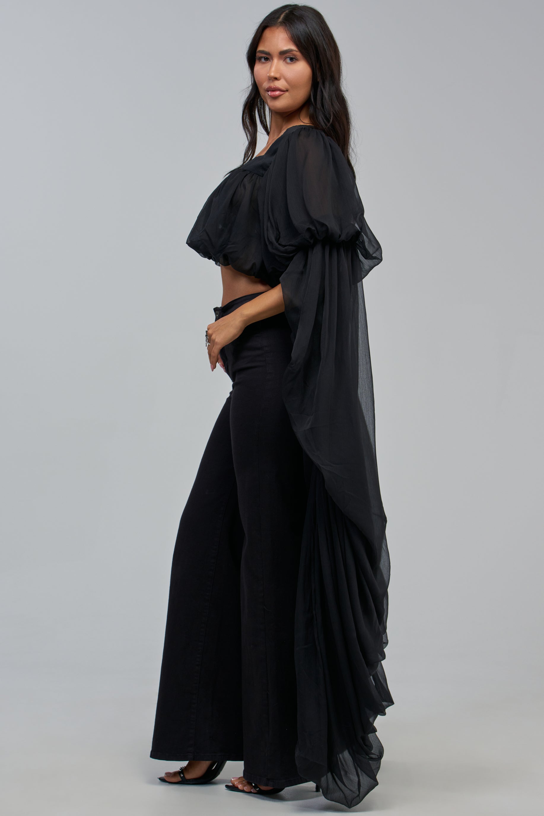 A woman stands in profile wearing the SLOUCHY SQUARE NECK LONG SLEEVE BLOUSE IN BLACK, featuring sheer black chiffon fabric and dramatic flowing sleeves. She pairs it with matching high-waisted flare pants, her long dark hair cascading down as she poses against a plain light gray background.