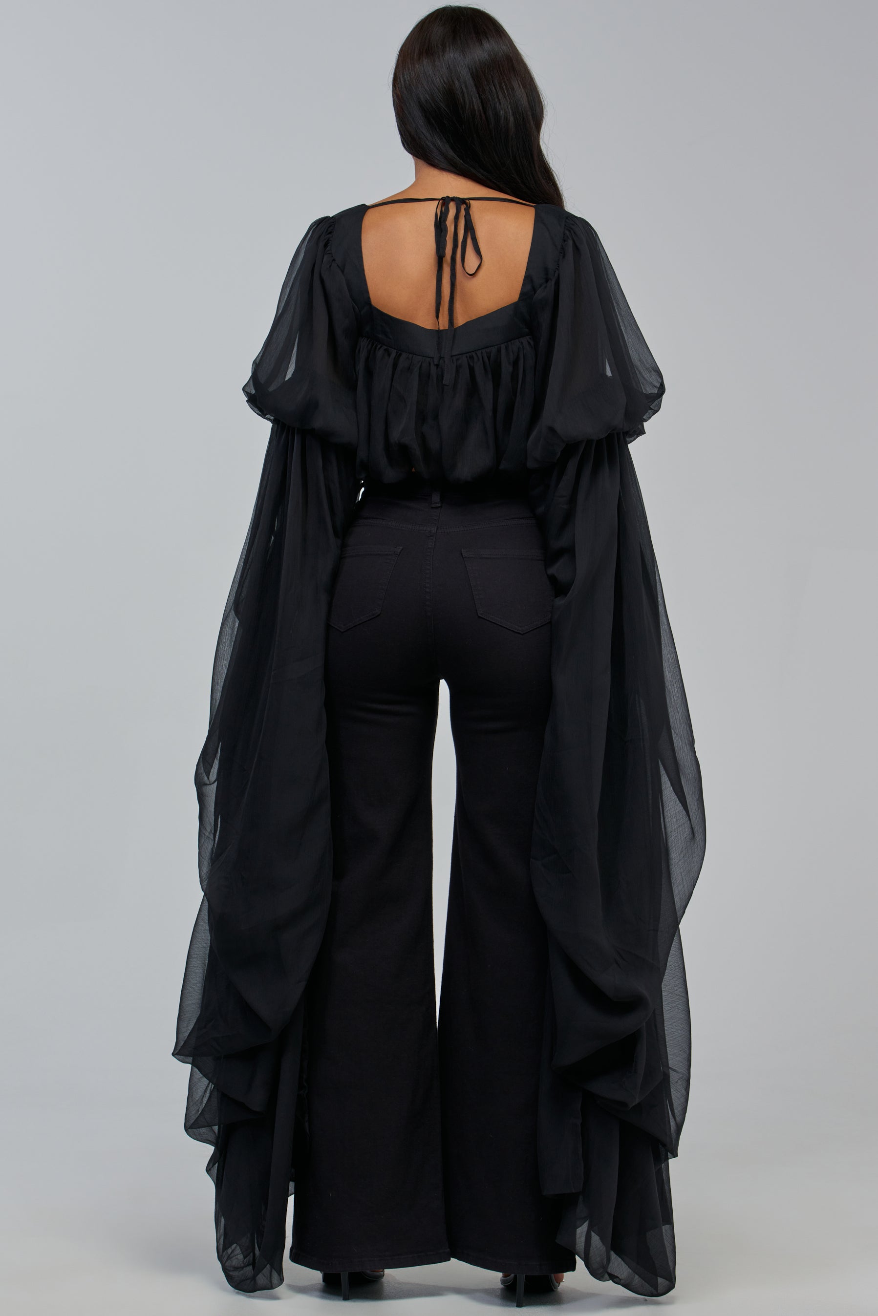 A person with long dark hair wears the SLOUCHY SQUARE NECK LONG SLEEVE BLOUSE IN BLACK, a dramatic and flowing chiffon piece with an open back, styled with high-waisted black pants and black shoes, standing facing away against a plain background.