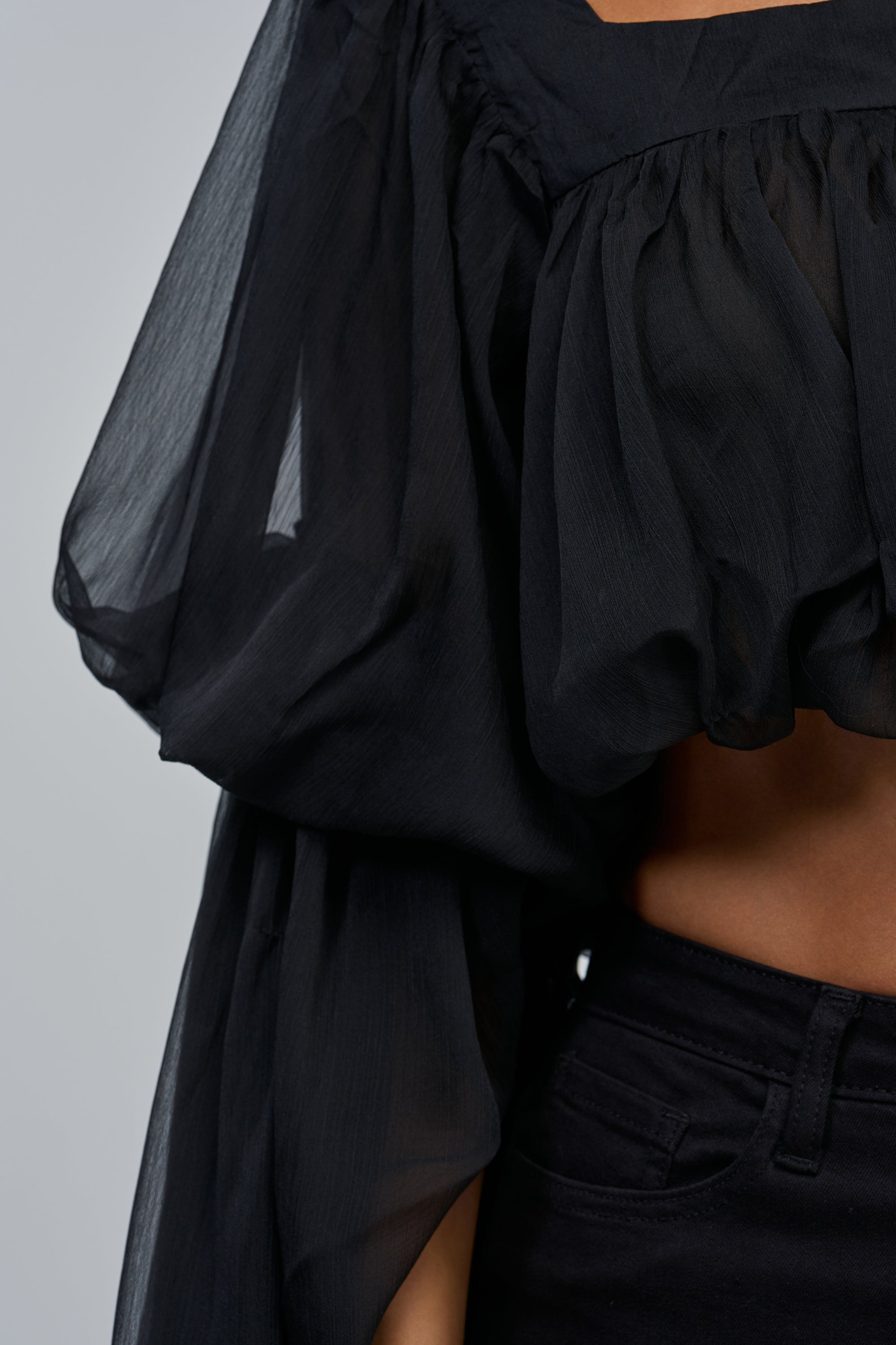 A close-up of a person wearing the SLOUCHY SQUARE NECK LONG SLEEVE BLOUSE IN BLACK, a sheer chiffon, off-the-shoulder top with voluminous long sleeves, paired with high-waisted black pants and revealing a hint of midriff for a boho chic look.
