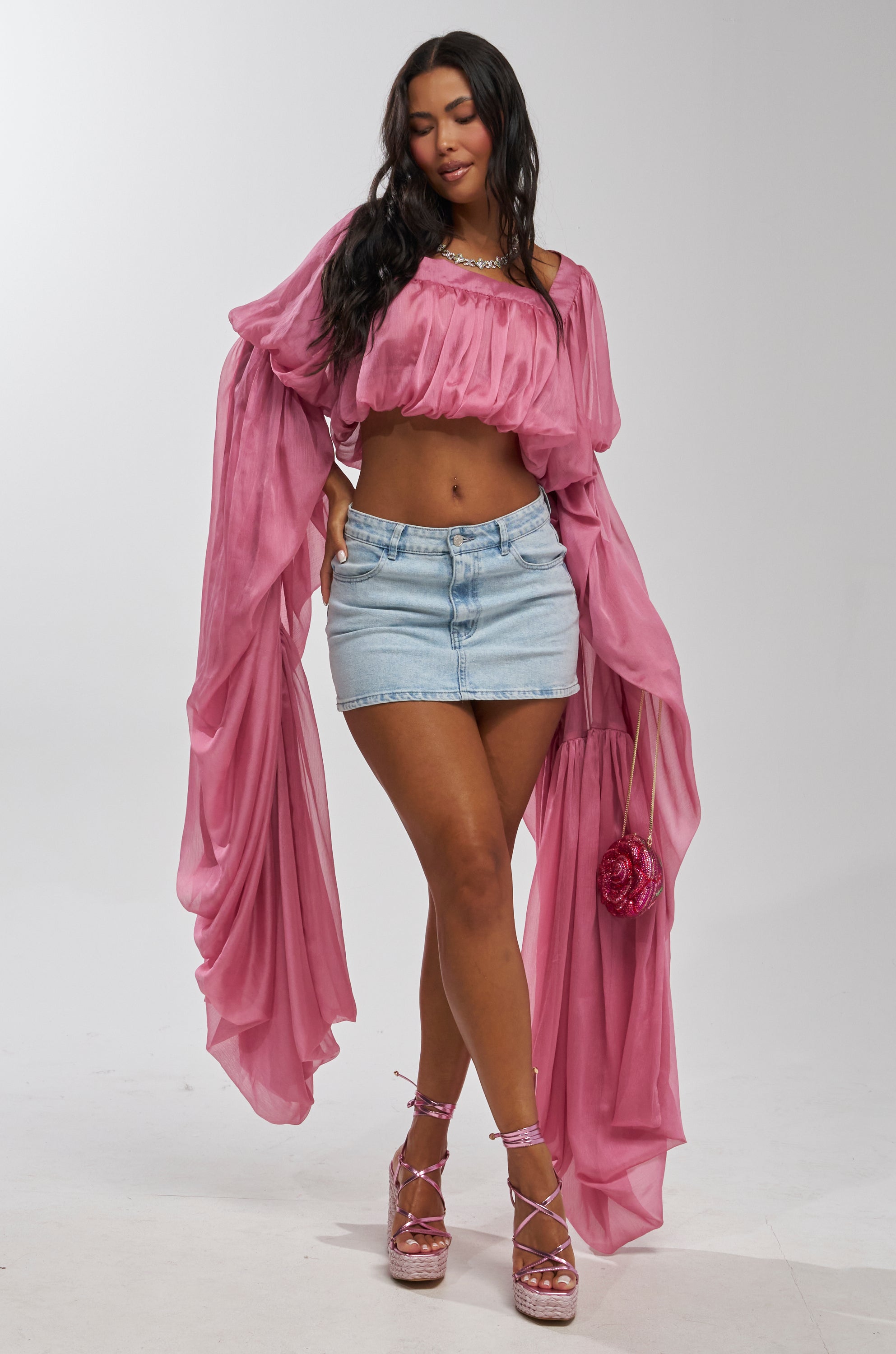 A woman poses in the SLOUCHY SQUARE NECK LONG SLEEVE BLOUSE TOP IN MAUVE, paired with a light blue denim mini skirt, pink strappy platform heels, and a small pink flower-shaped handbag against a plain white background.
