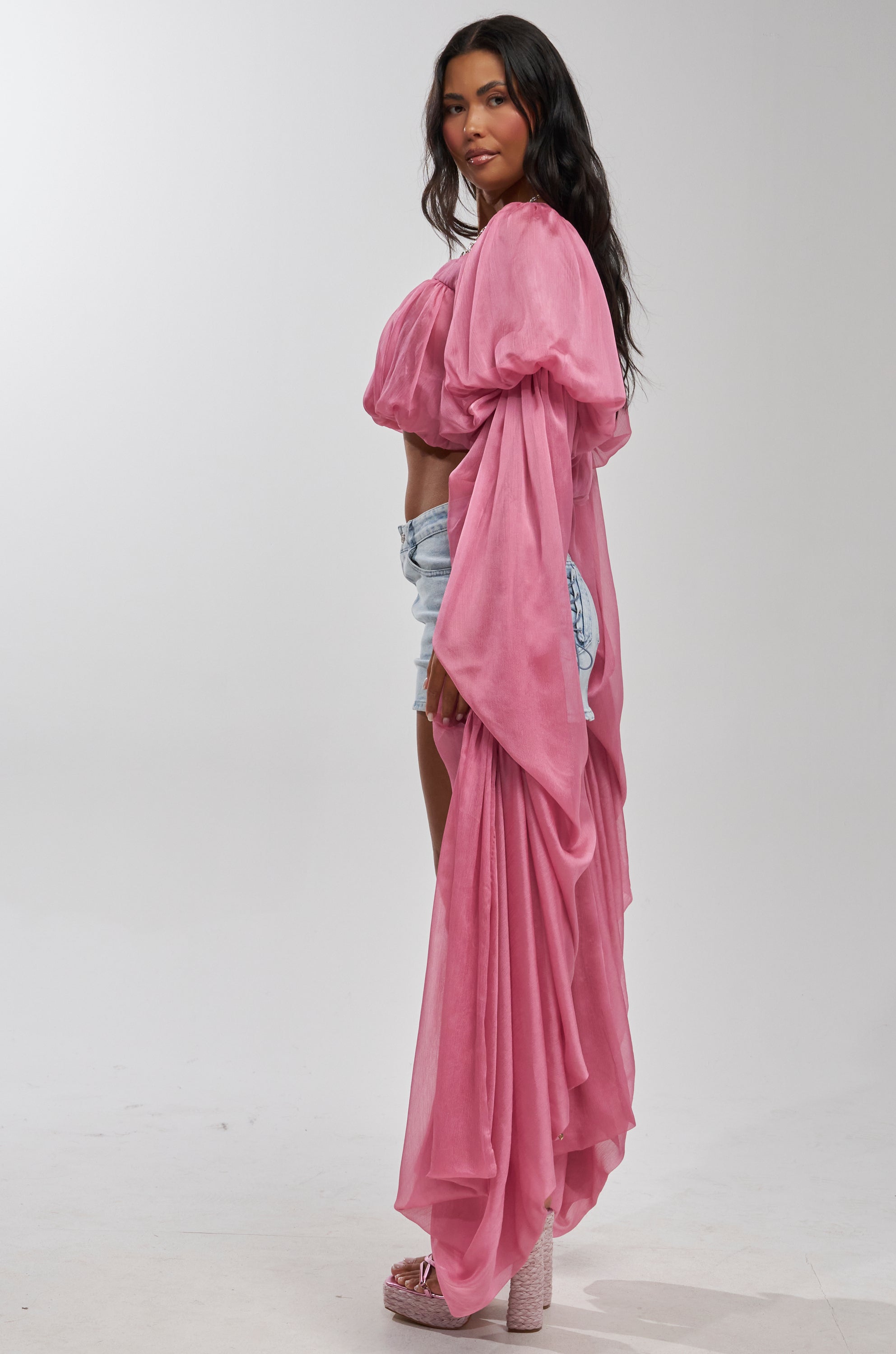 A woman in profile wears the SLOUCHY SQUARE NECK LONG SLEEVE BLOUSE TOP IN MAUVE over a cropped top and light denim shorts, paired with pink platform heels, against a plain white background.
