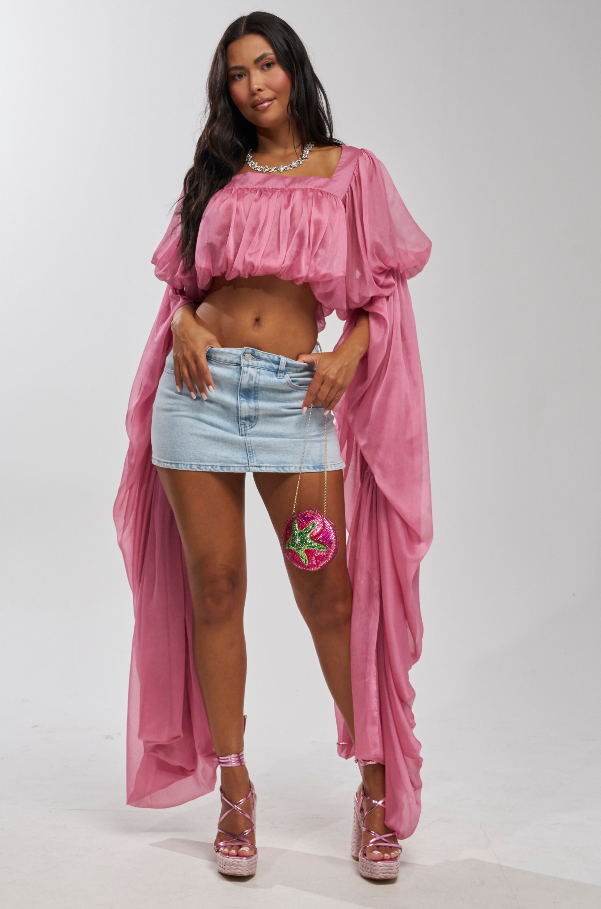 A woman poses in a SLOUCHY SQUARE NECK LONG SLEEVE BLOUSE TOP IN MAUVE, paired with a light blue denim mini skirt, platform pink heels, a necklace, and holds a small glittery pink-and-green purse on a chain.