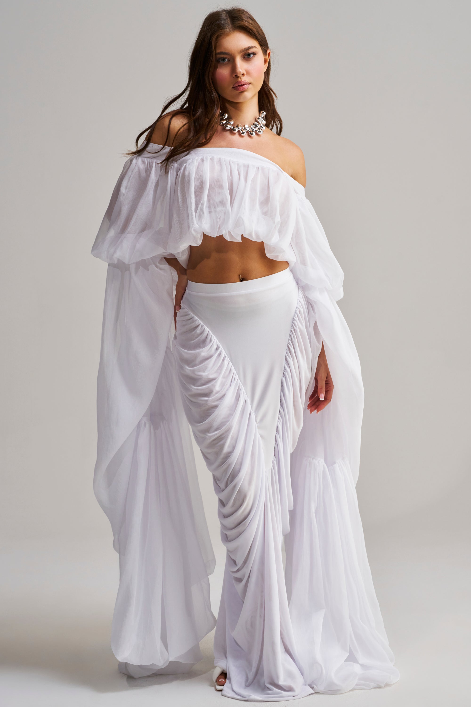 A woman poses against a plain light background, wearing the SLOUCHY SQUARE NECK LONG SLEEVE BLOUSE TOP IN WHITE with an off-the-shoulder neckline, paired with a matching ruched skirt and a statement necklace.