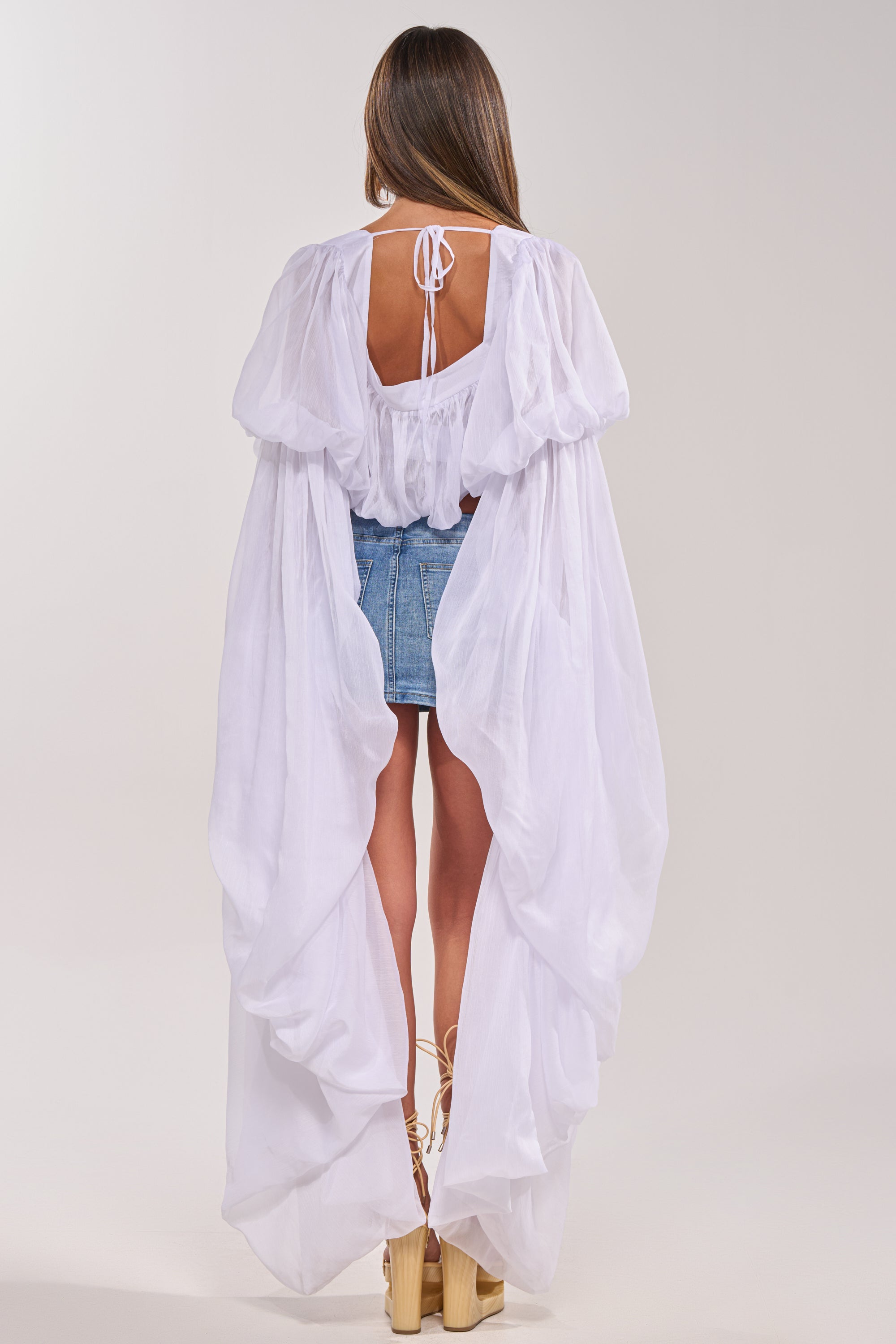 A woman faces away, wearing the SLOUCHY SQUARE NECK LONG SLEEVE BLOUSE TOP IN WHITE under a sheer white cape, paired with a short denim skirt and platform heels. Her straight hair is loose, and the open-back cape ties at the neck.