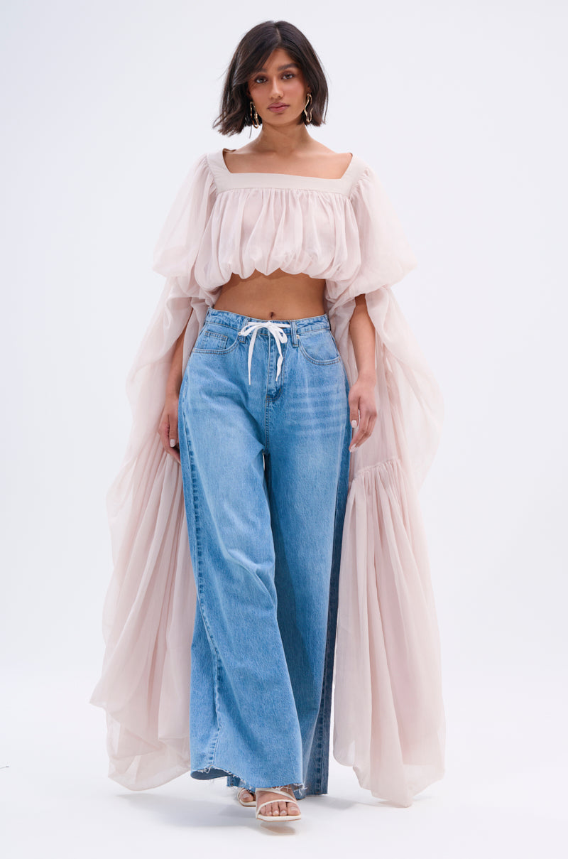 A woman walks confidently against a plain white background, wearing the SLOUCHY SQUARE NECK LONG SLEEVE BLOUSE TOP in pale pink with wide-leg blue jeans and white heeled sandals.