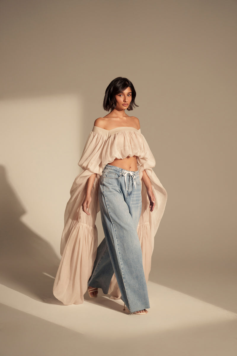 A woman stands in soft light wearing a SLOUCHY SQUARE NECK LONG SLEEVE BLOUSE TOP with billowy sleeves and wide-leg blue jeans. She is barefoot, looking forward against a neutral background.