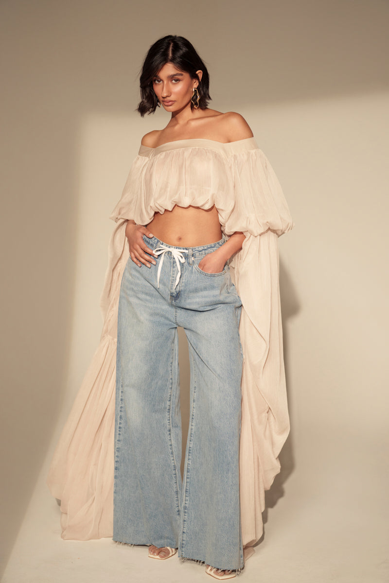 A woman poses confidently in the SLOUCHY SQUARE NECK LONG SLEEVE BLOUSE TOP paired with high-waisted, wide-leg jeans and heeled sandals, standing against a beige background.