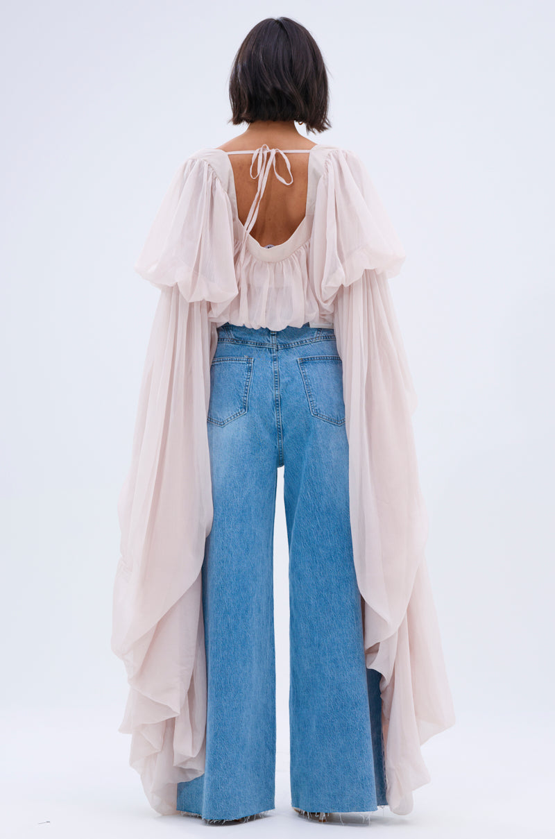 A person with short dark hair, facing away, wears wide-leg blue jeans and the SLOUCHY SQUARE NECK LONG SLEEVE BLOUSE TOP—flowy and sheer with billowy sleeves and an open back tied with a string—against a plain, light background.