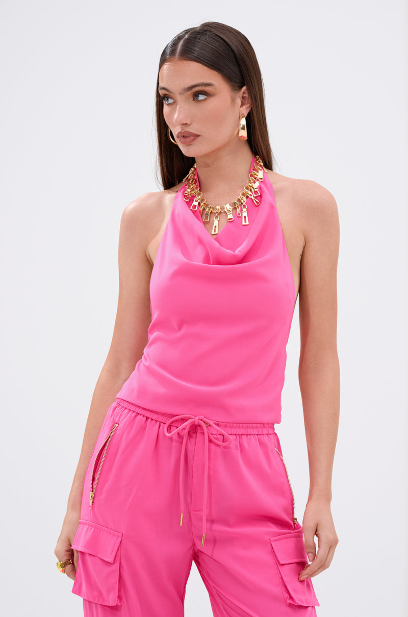 A woman in the UP TO NO GOOD HALTER BLOUSE IN PINK, featuring an open-back design, paired with matching cargo pants and gold accessories, poses against a white background.