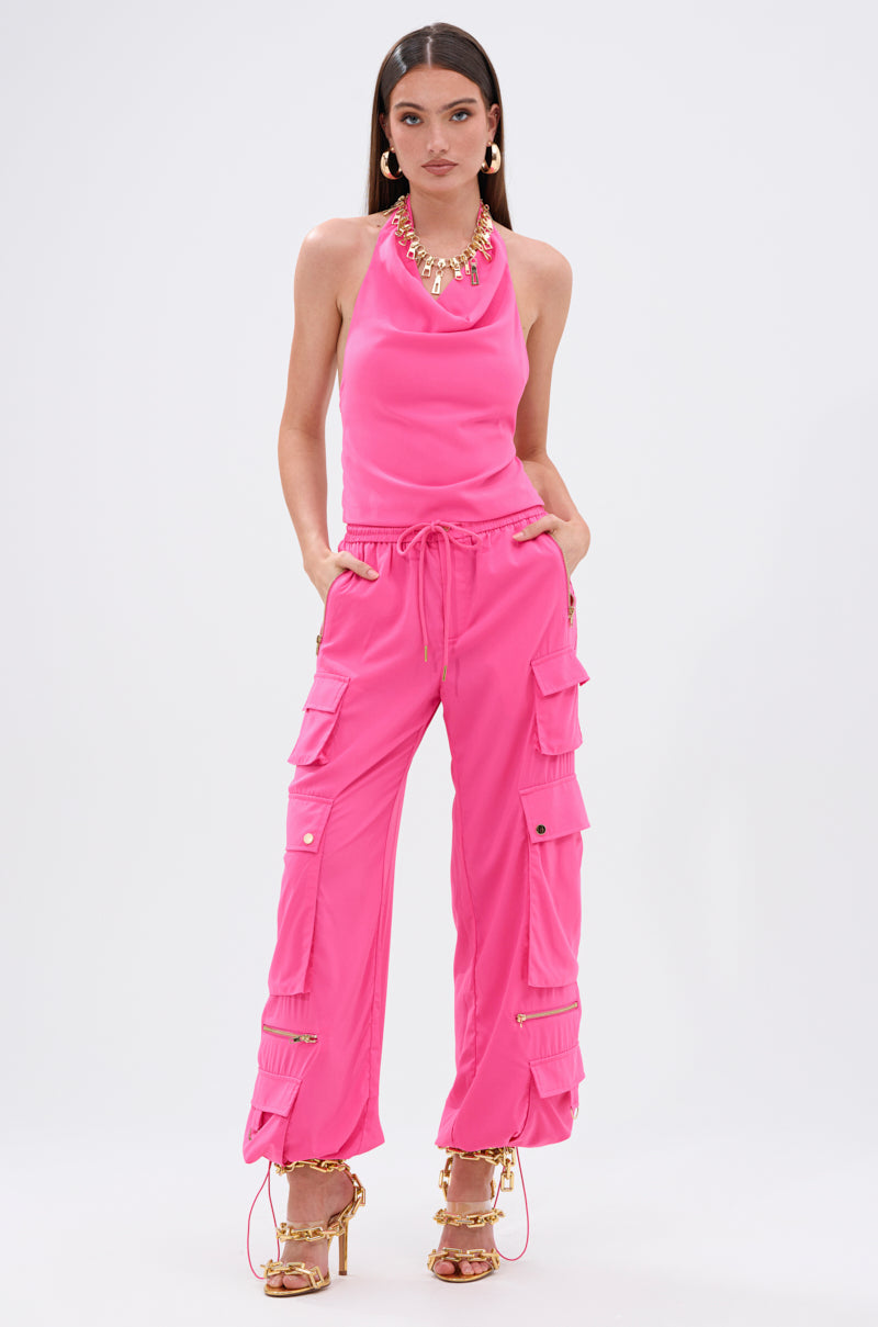 A woman stands against a white background wearing the UP TO NO GOOD HALTER BLOUSE IN PINK with matching pink cargo pants, gold jewelry, and strappy high heels. Hands in her pockets, she looks confidently at the camera.