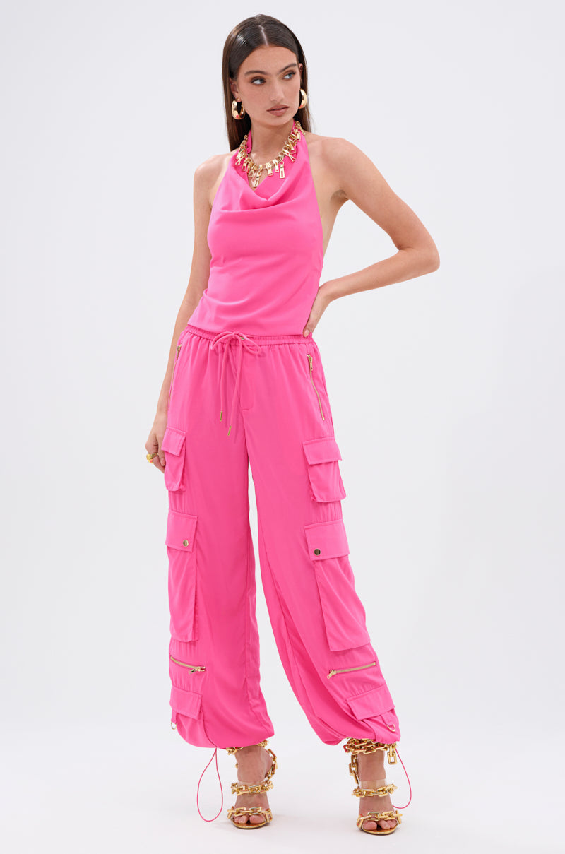 A woman models the UP TO NO GOOD HALTER BLOUSE IN PINK, a vibrant open-back top paired with matching cargo pants with pockets and drawstring hems. She accessorizes with gold jewelry and pink heels featuring gold chain details against a plain white background.