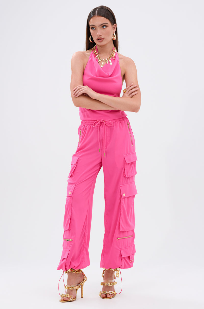 A woman wears the UP TO NO GOOD HALTER BLOUSE IN PINK with a cowl neckline and matching pink cargo pants featuring pockets and gold buttons, styled with gold jewelry and strappy high-heeled sandals against a plain background.