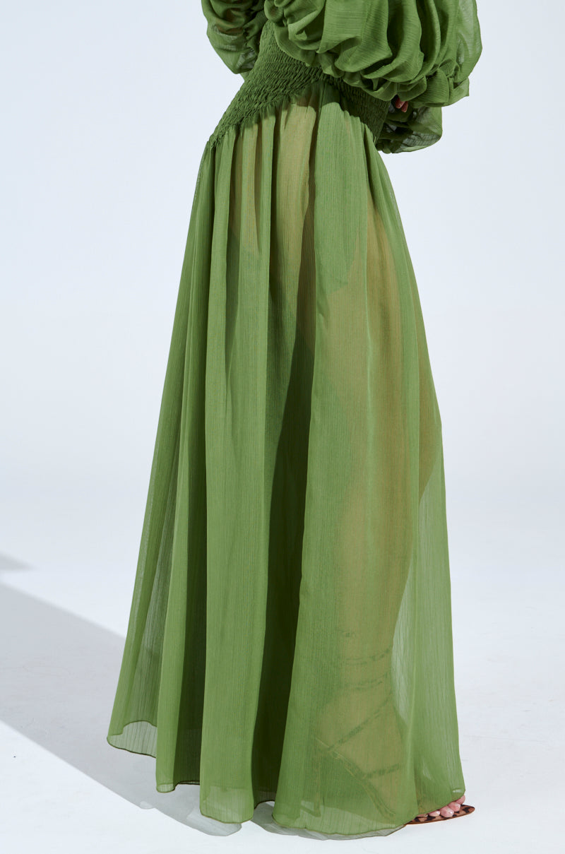 A person wears the LAURA RUCHED PALAZZO PANT IN GREEN, elegantly styled with a voluminous long dress effect and cinched at the waist to evoke a wide-leg silhouette. The sheer fabric subtly reveals the design beneath, all showcased against a plain studio backdrop for an ideal Spring look.