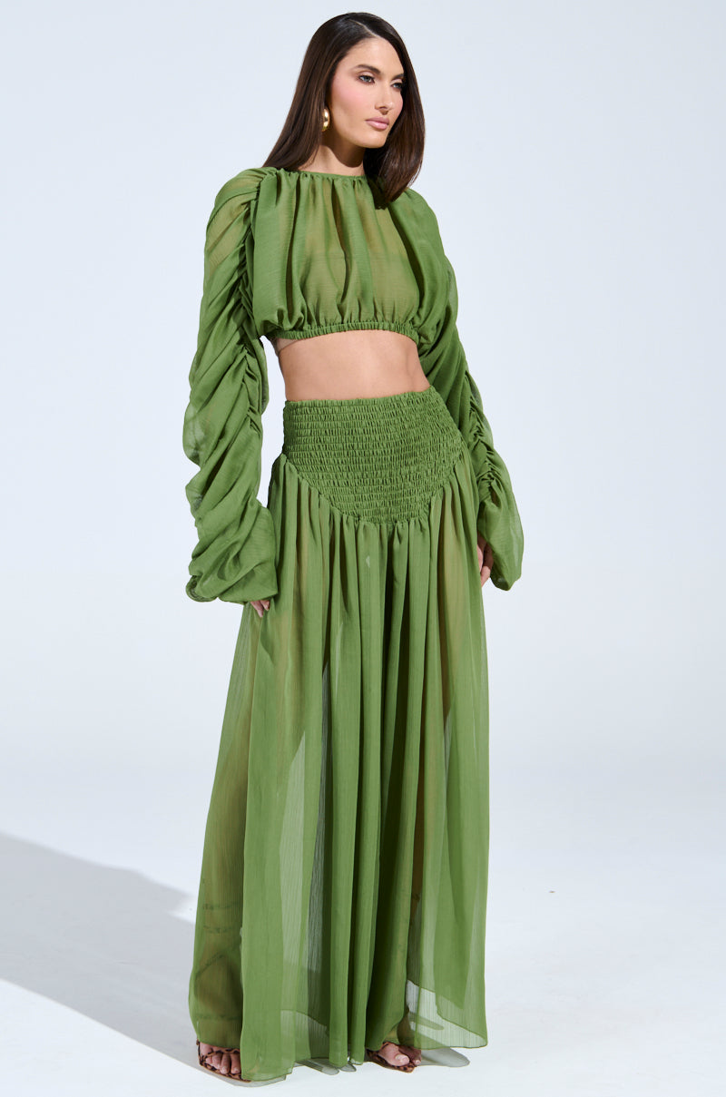 A woman stands against a white background, highlighting a spring staple. She wears a green, semi-sheer long-sleeve top with LAURA RUCHED PALAZZO PANT IN GREEN. The high-waisted, wide-leg pants beautifully complement her long hair.