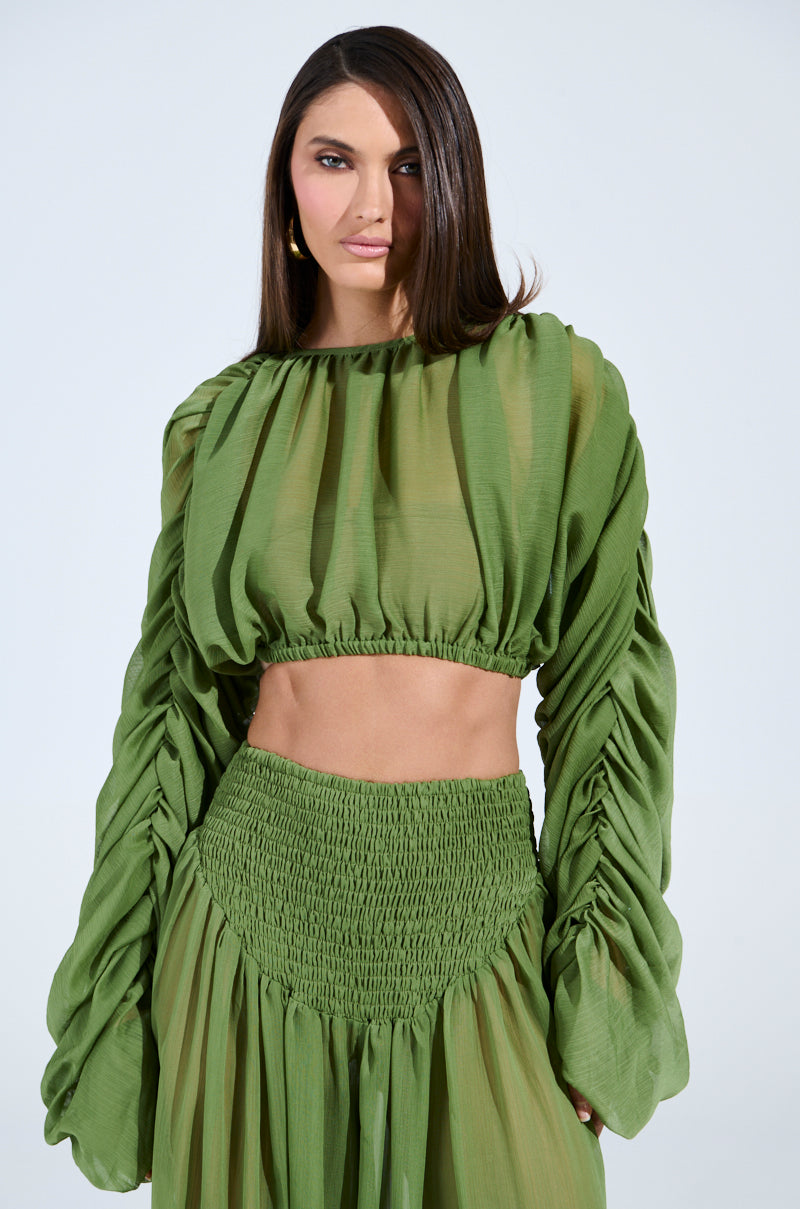 A woman wears a LAURA Ruched Cropped Blouse in vibrant green with a matching high-waisted skirt. Her straight dark hair and gold earrings add elegance against a simple, light backdrop.
