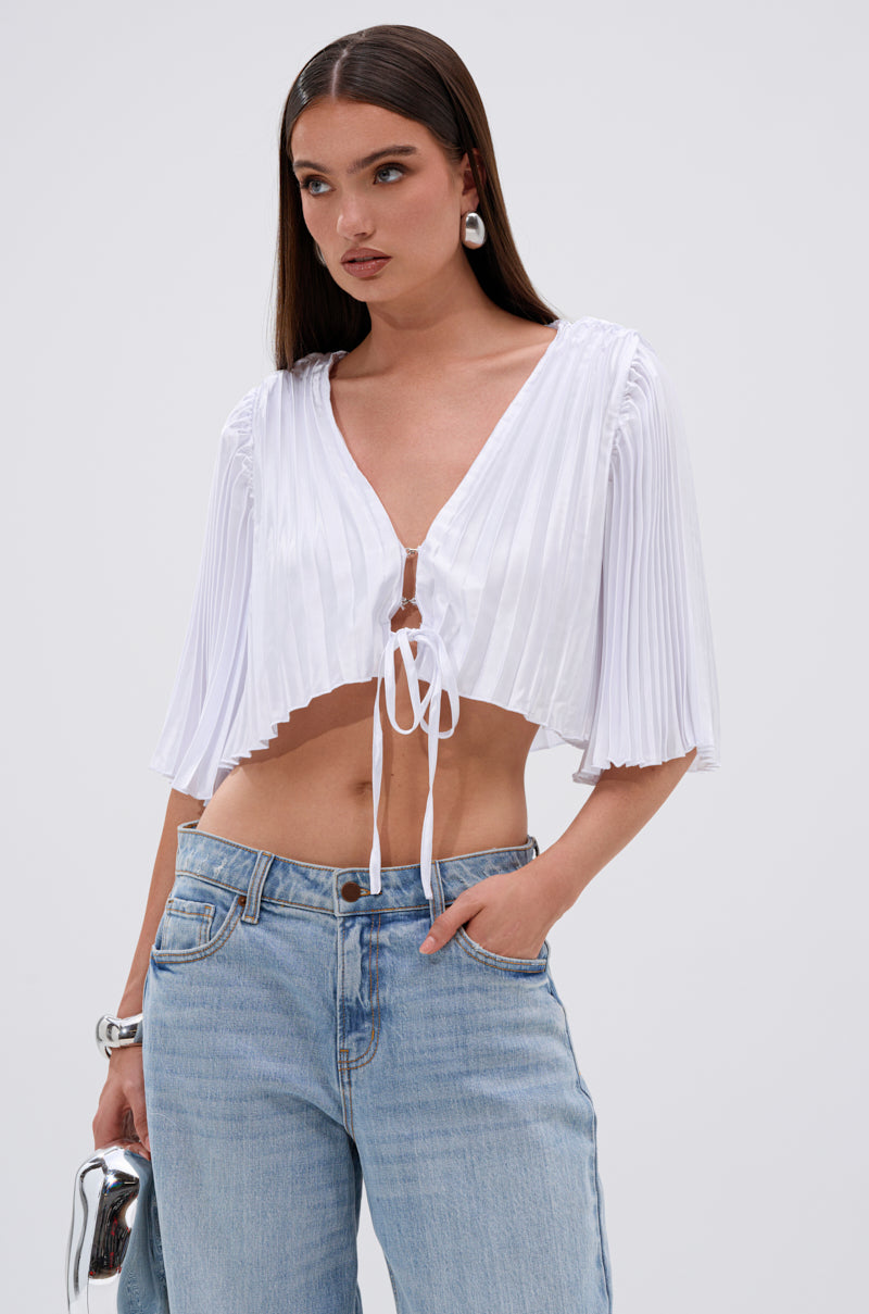 A woman stands against a plain background wearing the MARY LOO TIE FRONT BLOUSE—white, pleated, with elbow-length sleeves—and high-waisted light blue jeans, holding a silver clutch and looking to the side. 0
