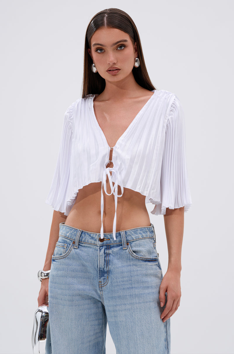A woman wears the MARY LOO TIE FRONT BLOUSE—white, pleated, with a deep V-neck—paired with light blue jeans. She has long straight hair, hoop earrings, and carries a small silver bag. 1