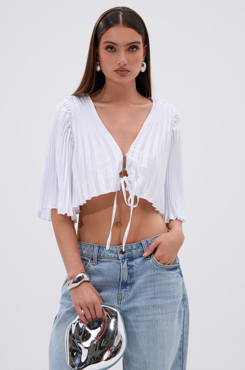 A woman with long brown hair wears the MARY LOO TIE FRONT BLOUSE—white, pleated, with elbow sleeves—paired with light blue jeans. She carries a shiny silver handbag and accessorizes with hoop earrings and a bracelet. 2