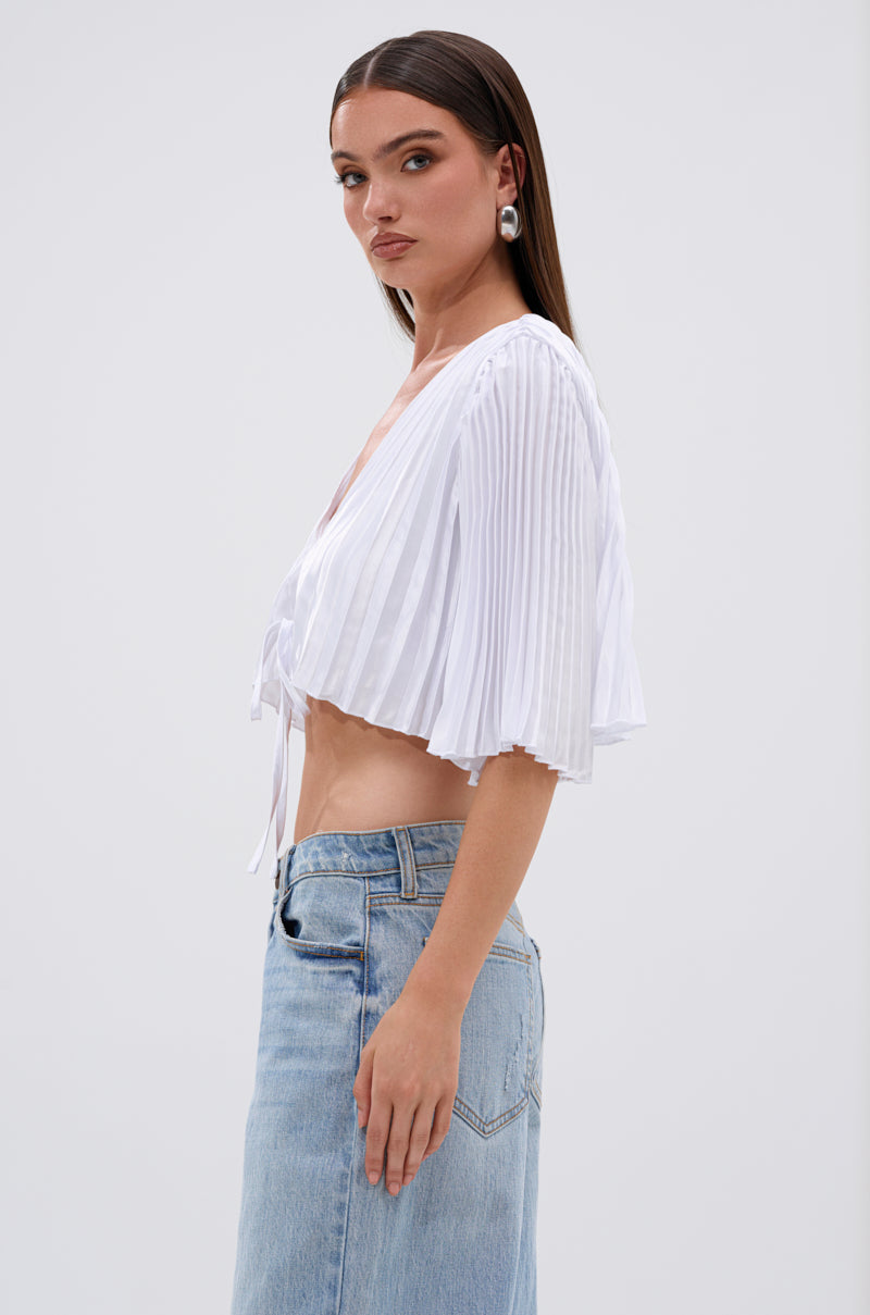A woman stands sideways against a plain background, wearing the MARY LOO TIE FRONT BLOUSE—white, pleated, with short sleeves—paired with light blue high-waisted jeans. She has long straight brown hair and large silver earrings. 3