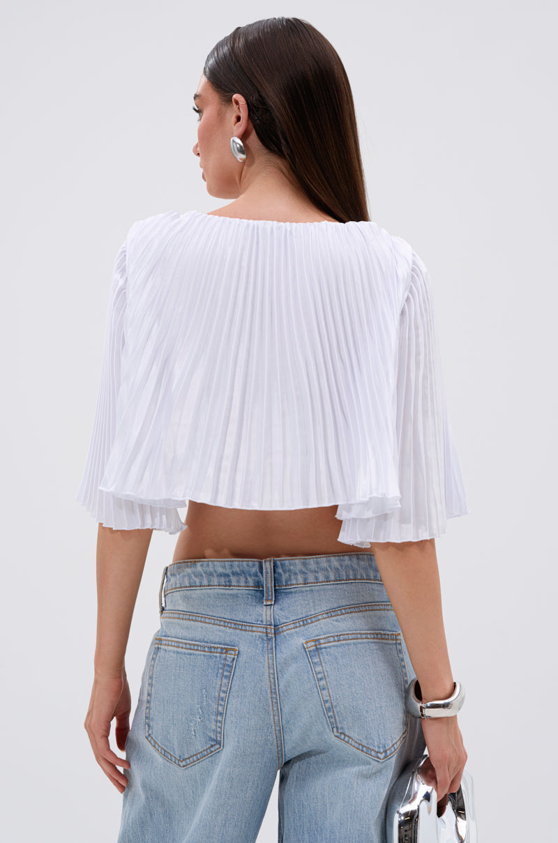 A woman stands facing away, wearing the MARY LOO TIE FRONT BLOUSE with wide sleeves and pleats, paired with light blue high-waisted jeans. She carries a metallic purse, wears chunky jewelry, and has straight brown hair down her back. 4
