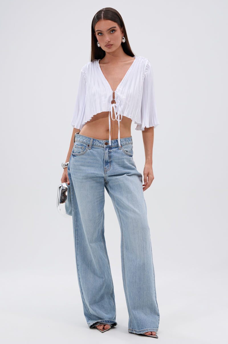 A woman stands against a plain background wearing the MARY LOO TIE FRONT BLOUSE—white, pleated, and wide-sleeved—paired with light blue high-waisted wide-leg jeans. She accessorizes with a silver handbag, hoop earrings, and heeled sandals. 5