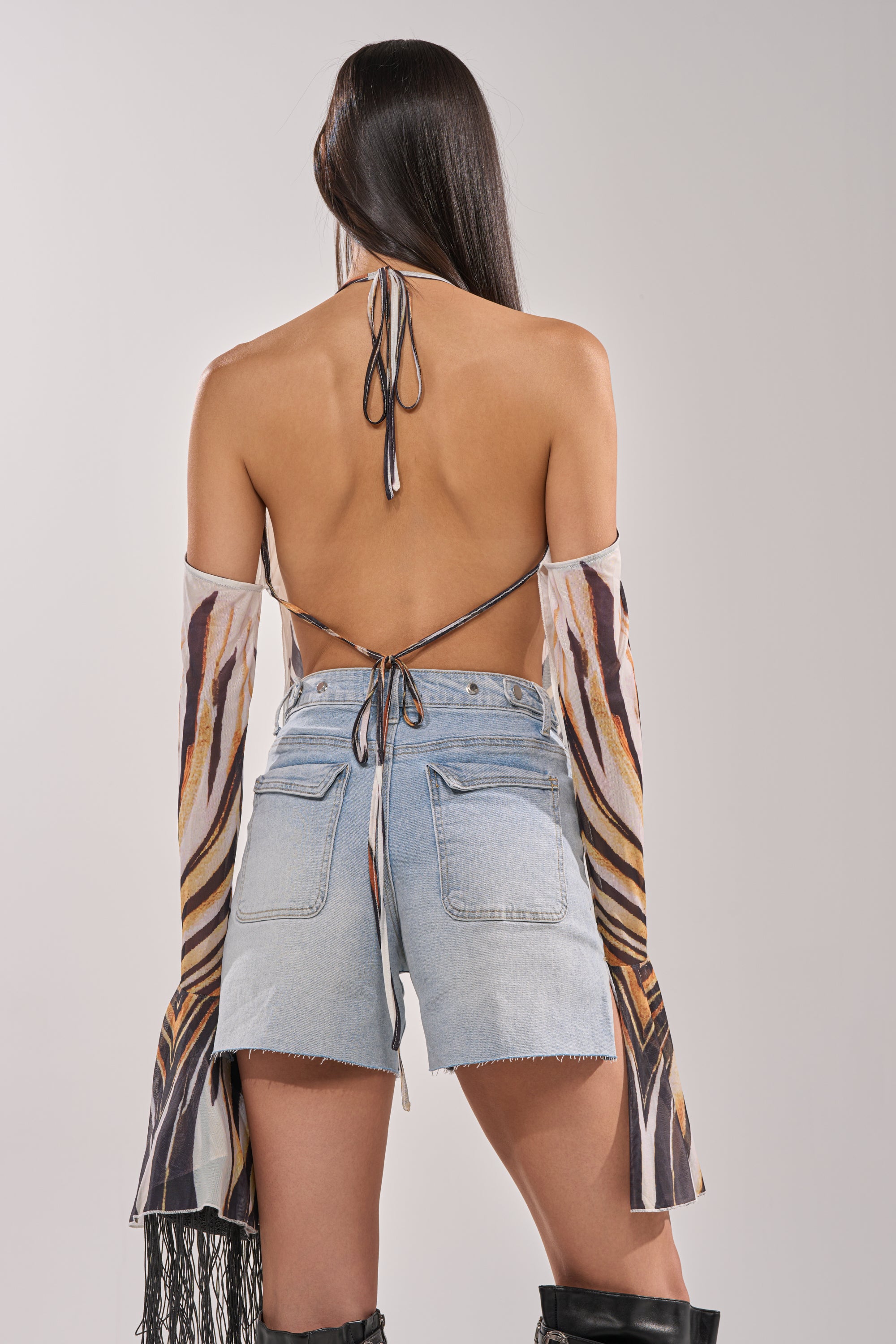 A woman with long dark hair is seen from behind, wearing the PRIMAL SPIRIT BLOUSE—a tiger print halter top with an open back—paired with light blue high-waisted denim shorts and black knee-high boots. 5